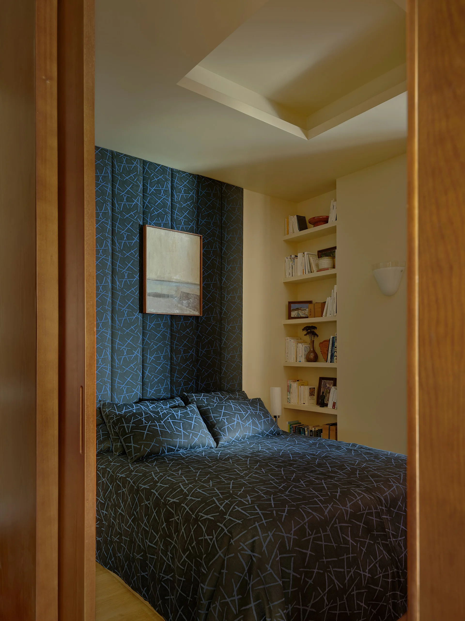 A bedroom viewed through a wooden doorway features a bed with matching navy patterned bedding and tall upholstered wall panels next to built-in shelves.