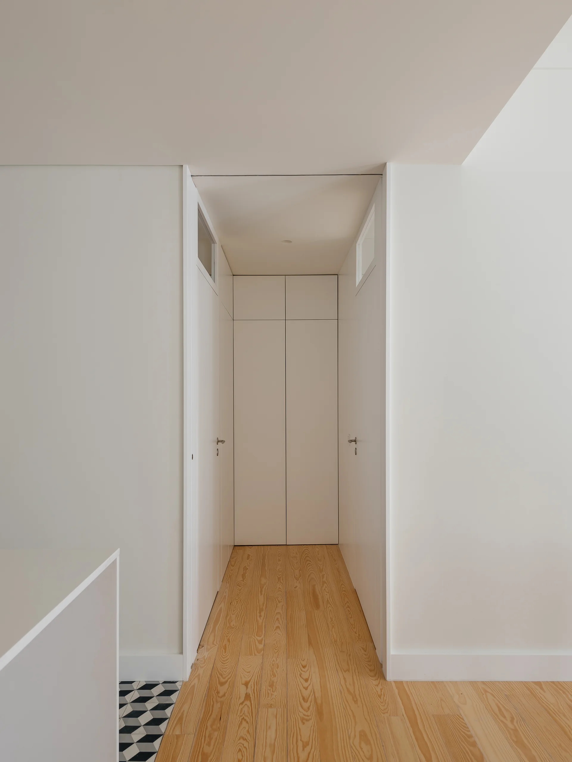 A narrow minimalist hallway features white flush-mount doors, transom windows, and light natural wood flooring.