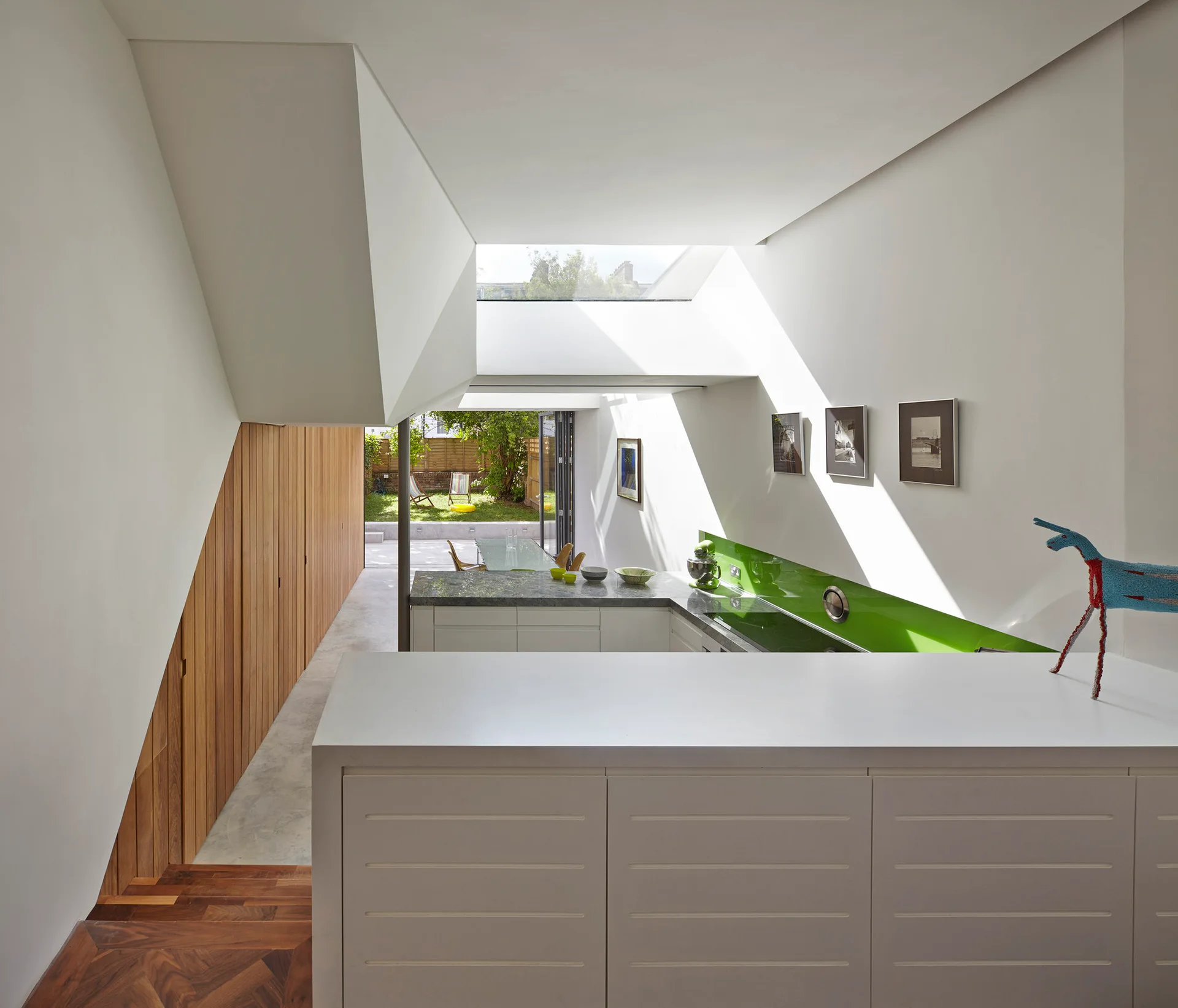 A modern white kitchen features a bright green glass backsplash, a large skylight overhead, and a slatted wood wall next to a staircase.