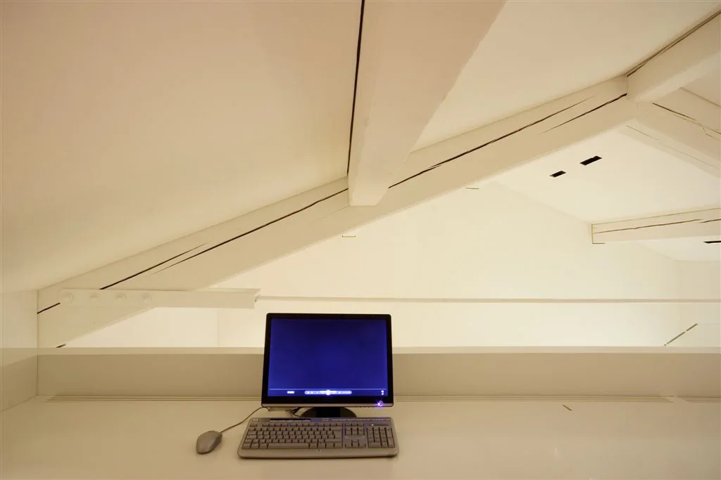 A minimalist computer workstation sits under a sloped attic ceiling with exposed white painted wooden beams.