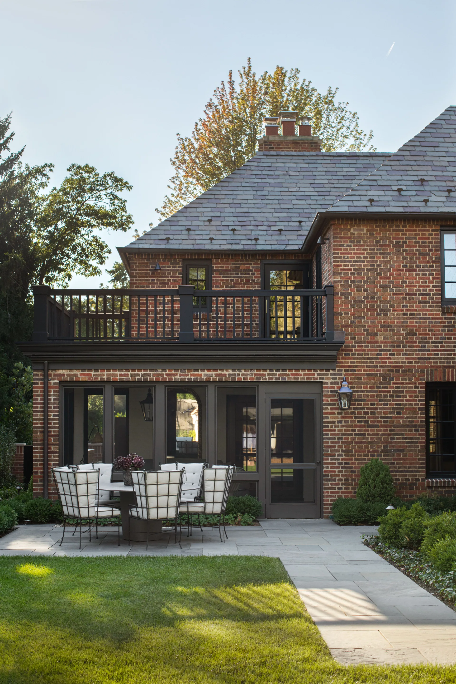 An outdoor stone patio features a round dining table with cushioned metal chairs set against a multi-story red brick home with a balcony.