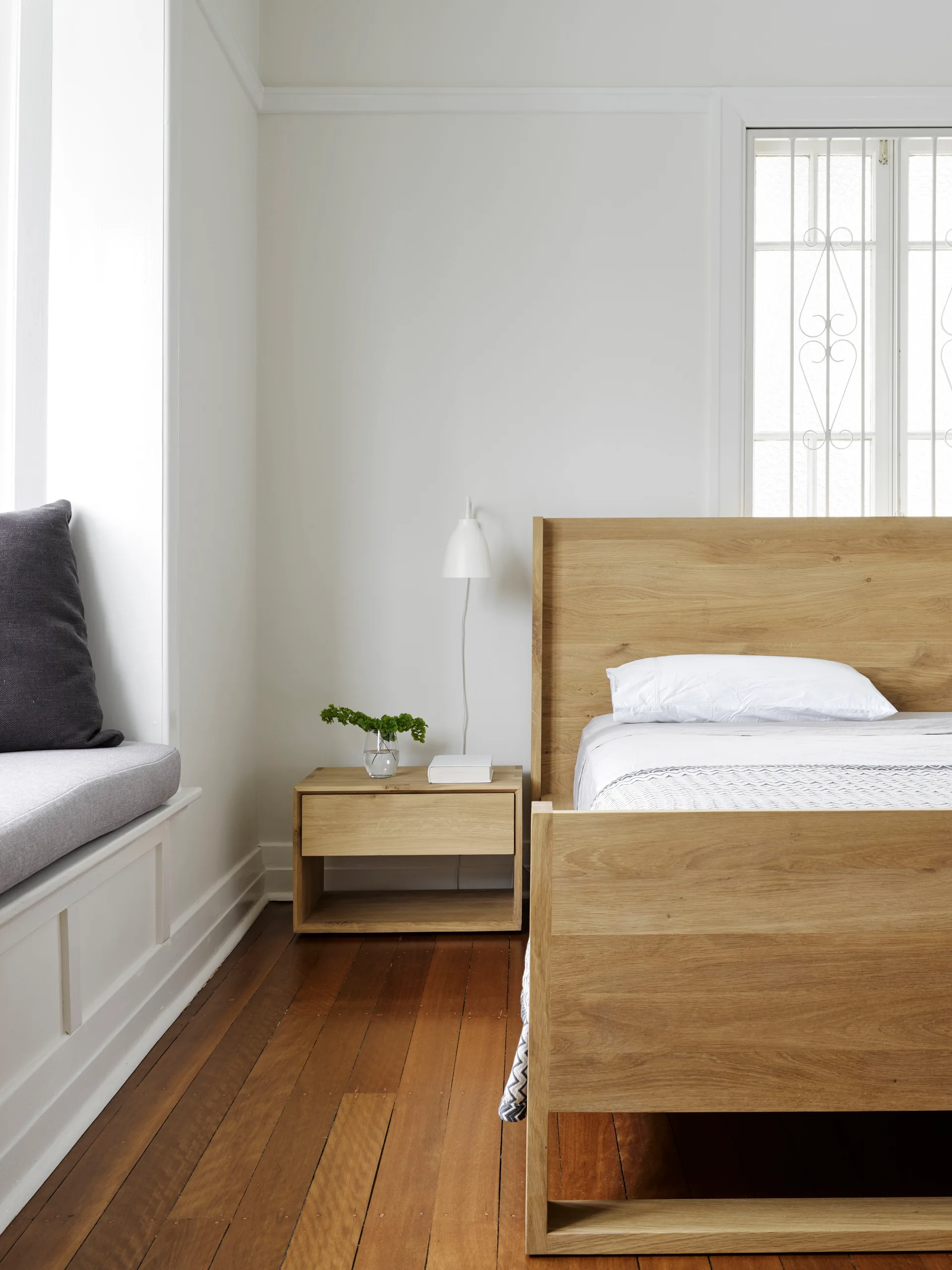 A minimalist bedroom featuring a light oak wood bed frame and a matching bedside drawer next to a built-in window seat with a grey cushion.