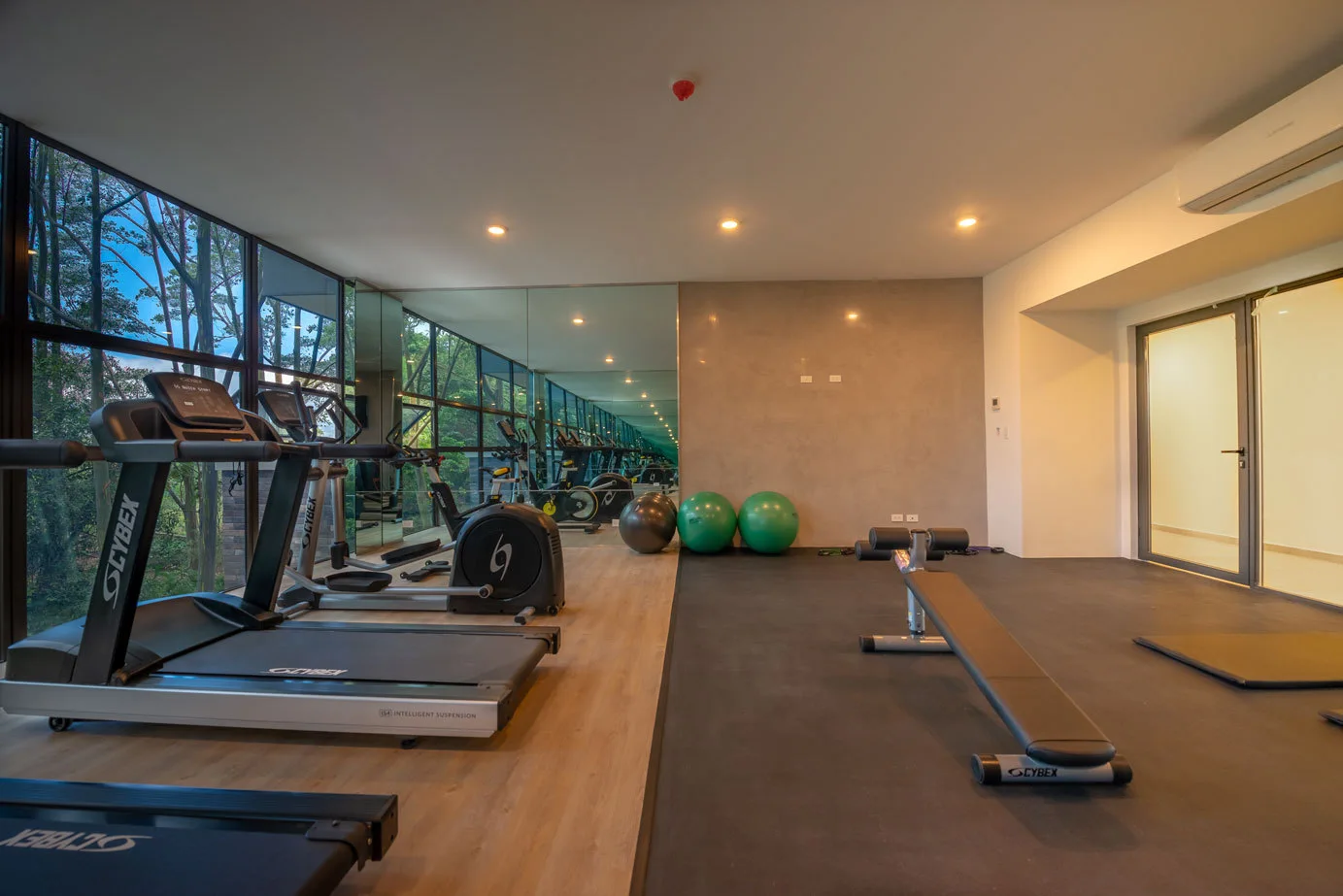 A modern fitness room features treadmills on light wood flooring, a weight bench on black rubber flooring, and a large mirrored wall.
