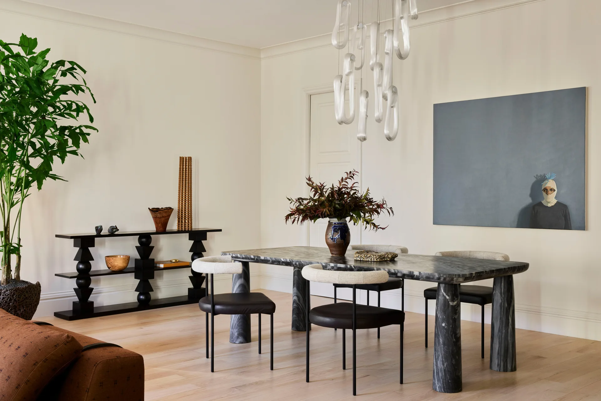 A modern dining room features a dark veined stone table with rounded chairs, a black tiered console, a large grey painting, and a sculptural pendant light.