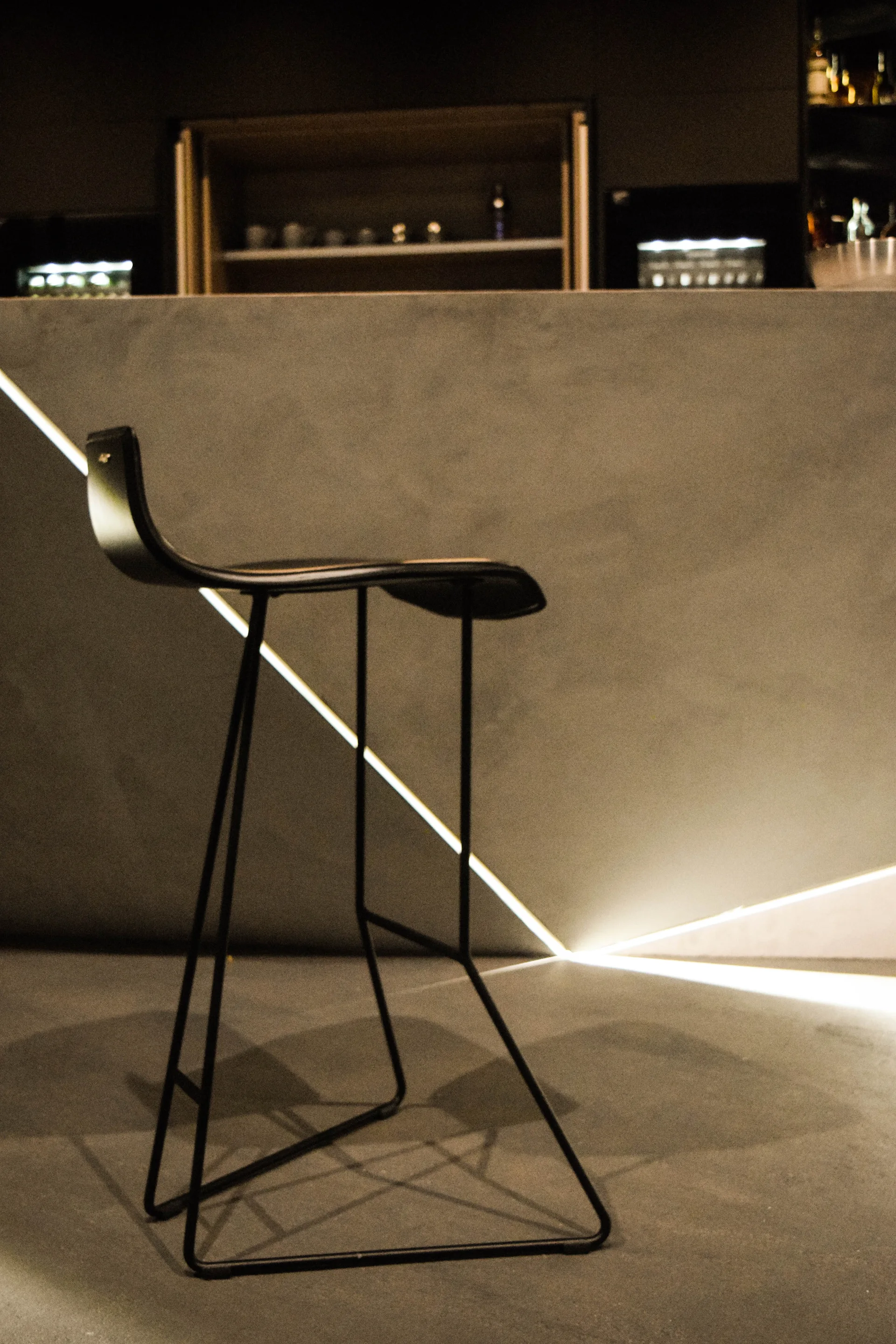 A slim black metal bar stool stands in front of a grey plaster bar counter accented by glowing diagonal LED light strips.