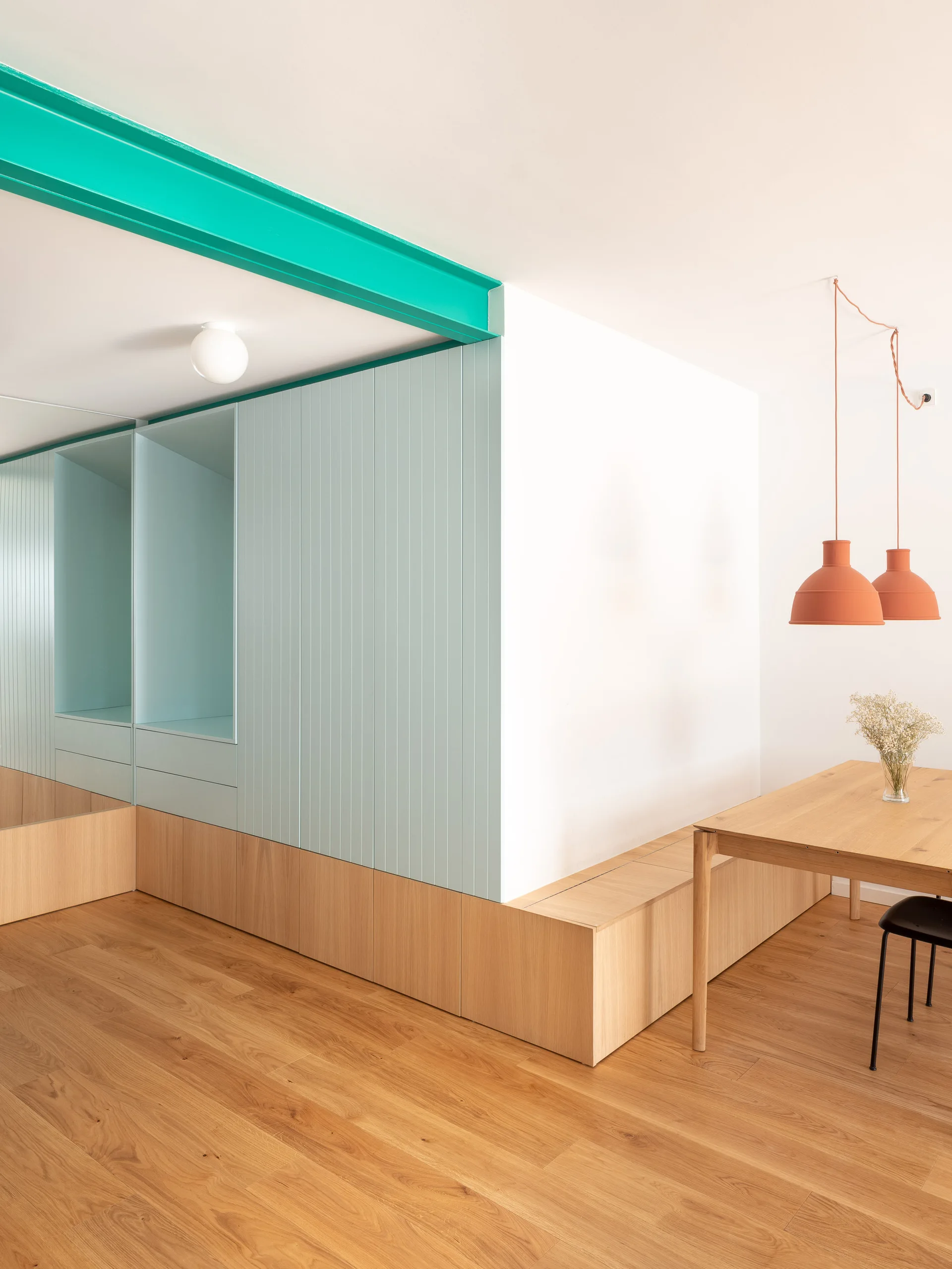 A modern dining room featuring a wooden table under orange pendant lights, pale blue built-in cabinets, and a bright turquoise ceiling beam.