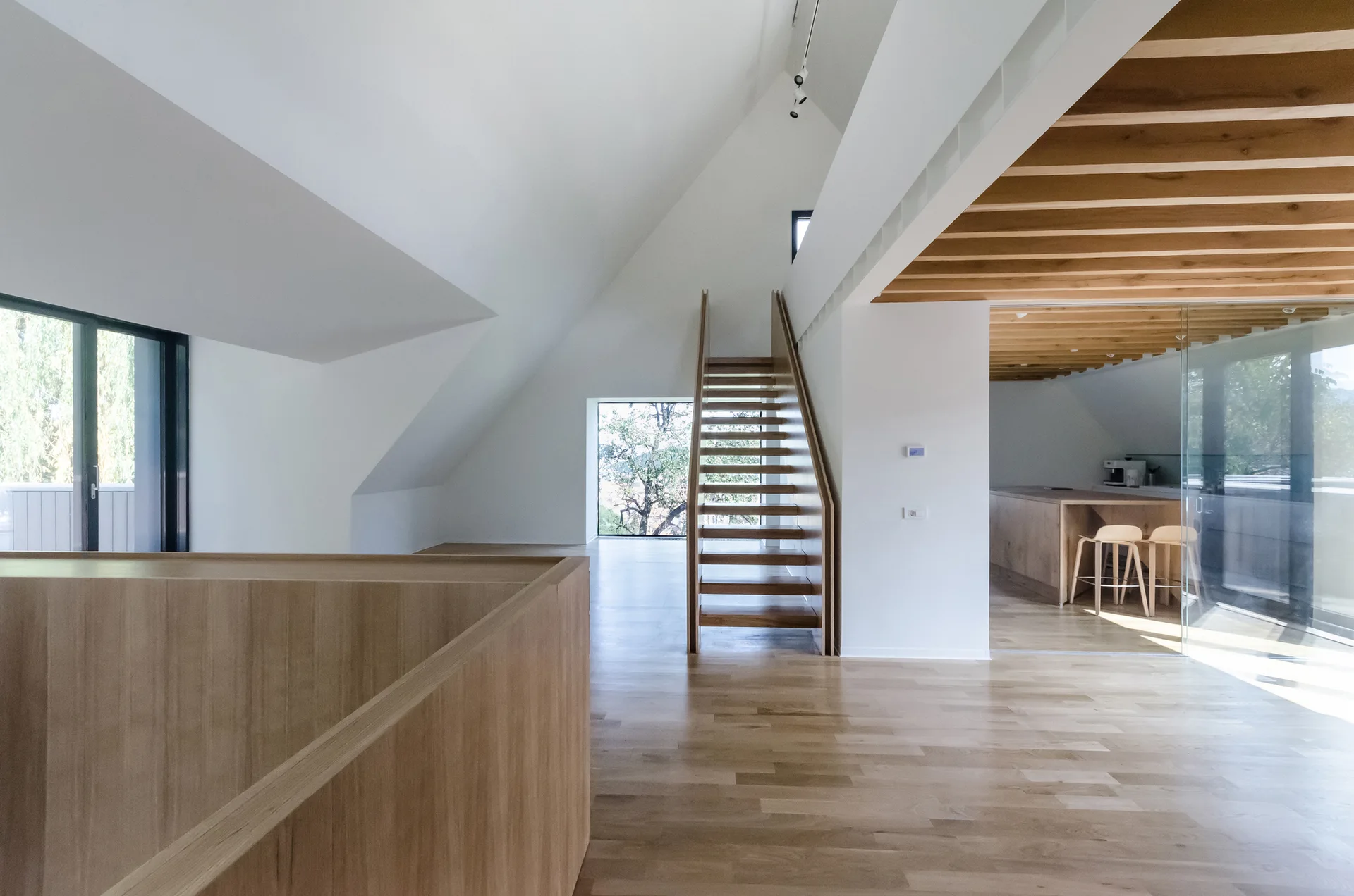 A minimalist interior features a central open-tread wood staircase, light wood floors, and white vaulted ceilings next to a glass-walled kitchen.