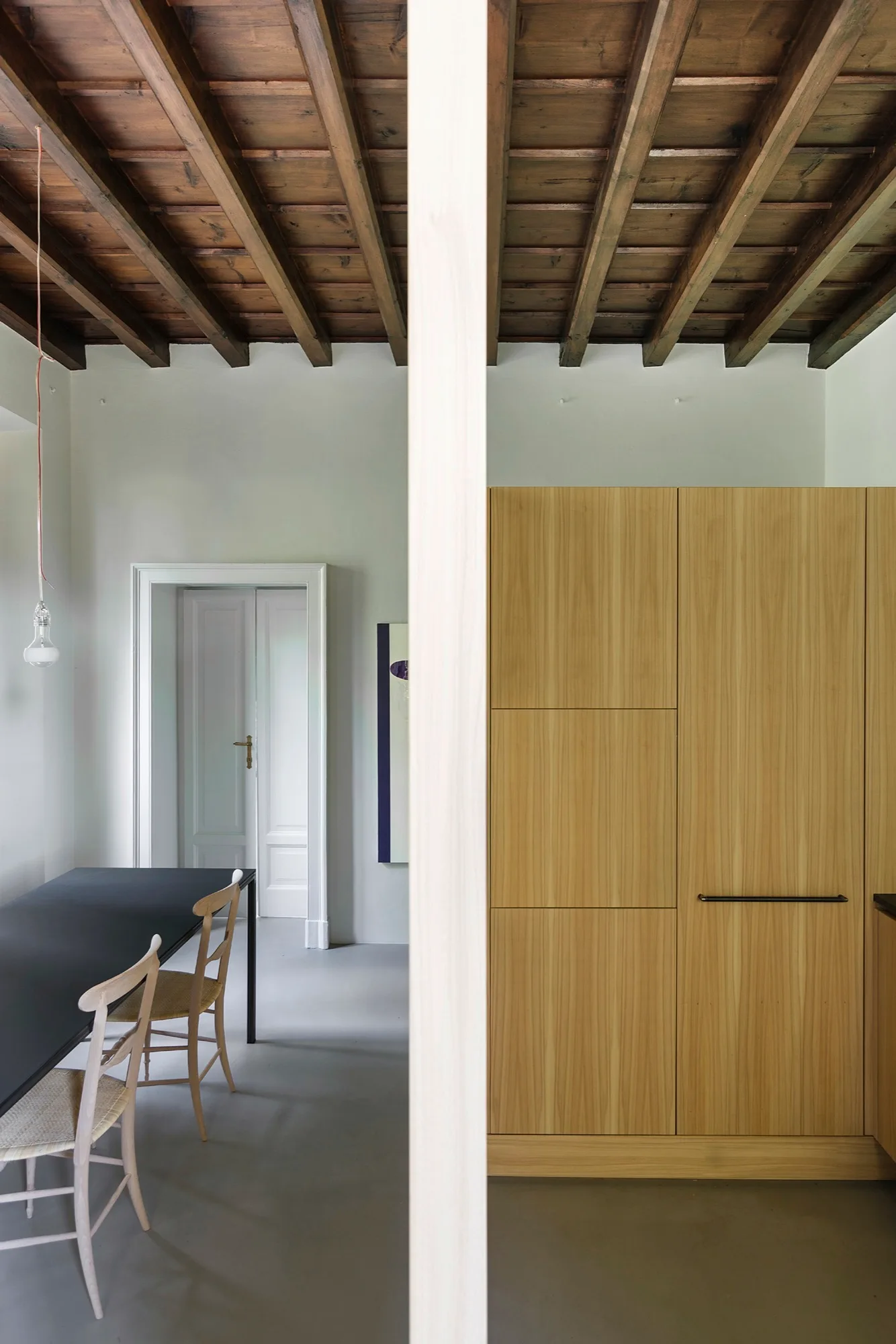 A minimalist dining space features a black table and light wood chairs set beneath an exposed dark wood beam ceiling and next to flat-panel cabinetry.