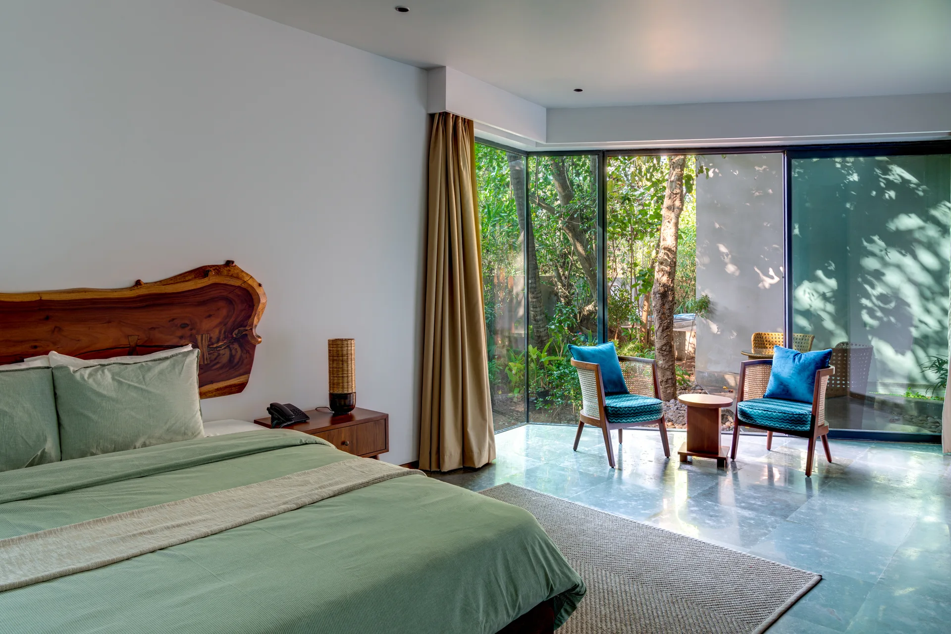 A modern bedroom features a bed with a live-edge wood headboard, sage green bedding, and a seating area with two teal chairs by a garden-view window.