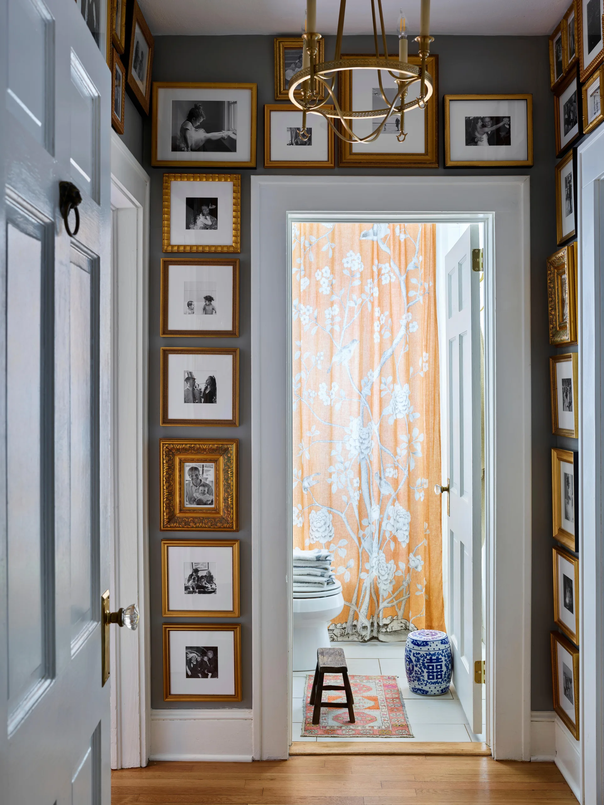 A narrow gray hallway with a dense gallery wall of gold-framed photos leads into a bathroom with an orange floral shower curtain.