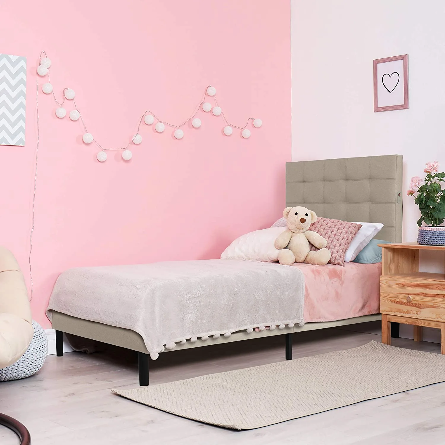 A kids bed with a beige tufted headboard stands against a pink wall decorated with white ball string lights next to a wooden nightstand.