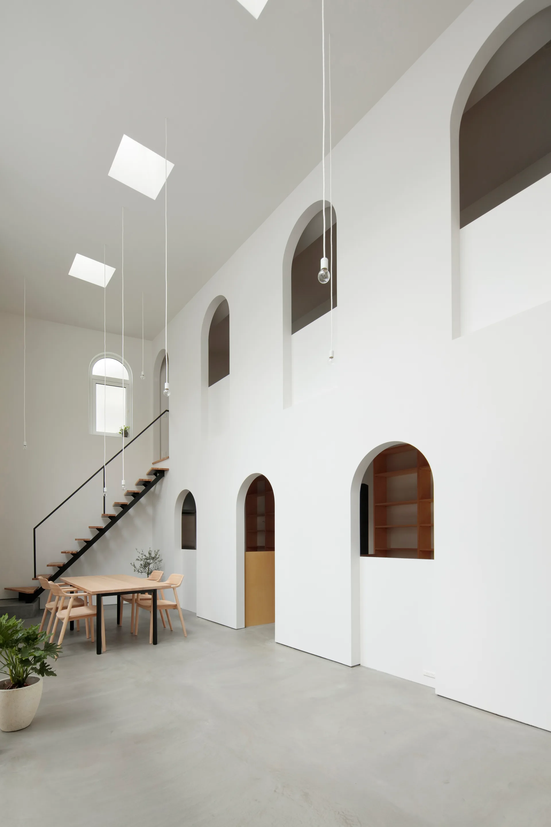 A minimalist dining room features a light wood table and chairs, a metal staircase, concrete floors, and a double-height white wall with arched openings.