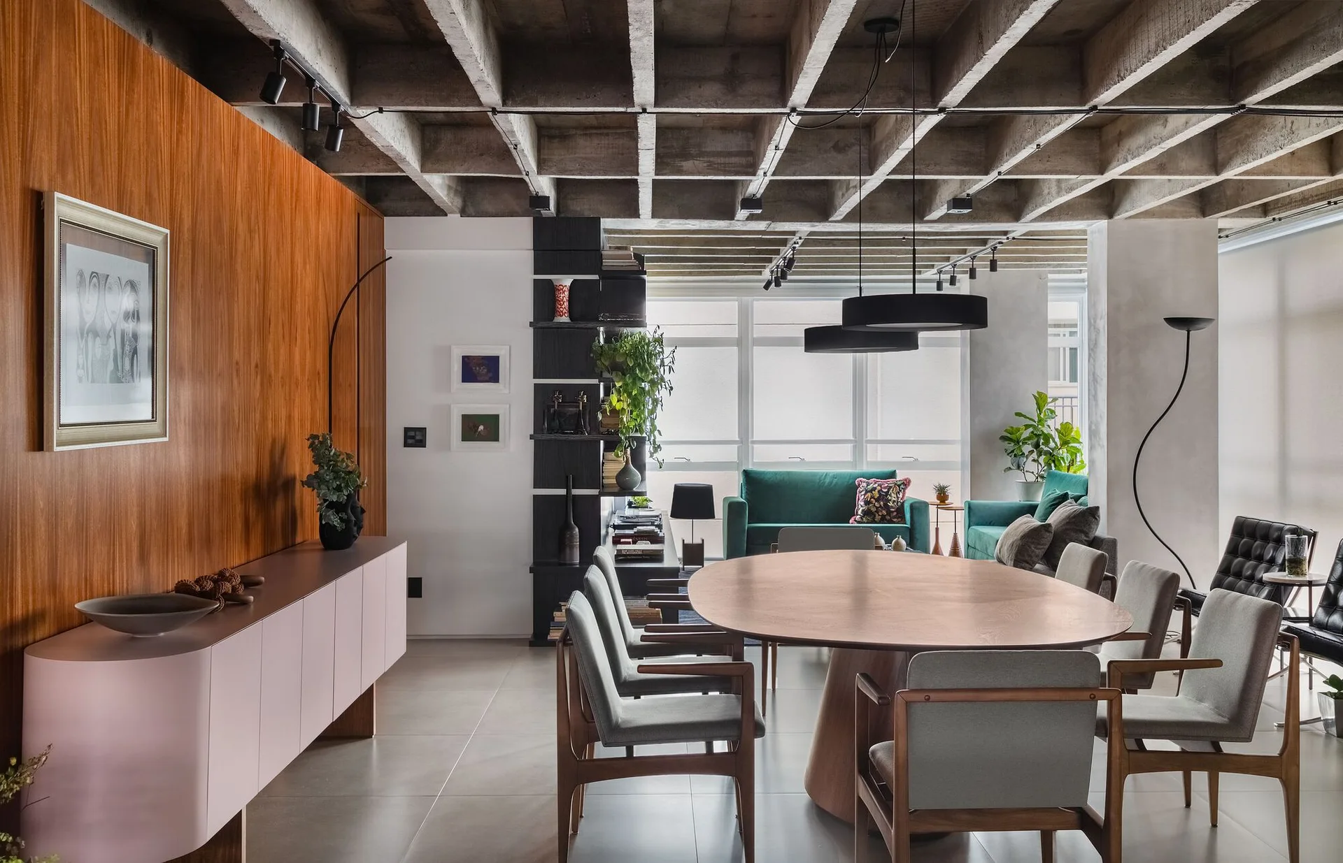 An open-plan dining room features a round wooden table, grey upholstered chairs, a pink sideboard, and an exposed concrete waffle ceiling.