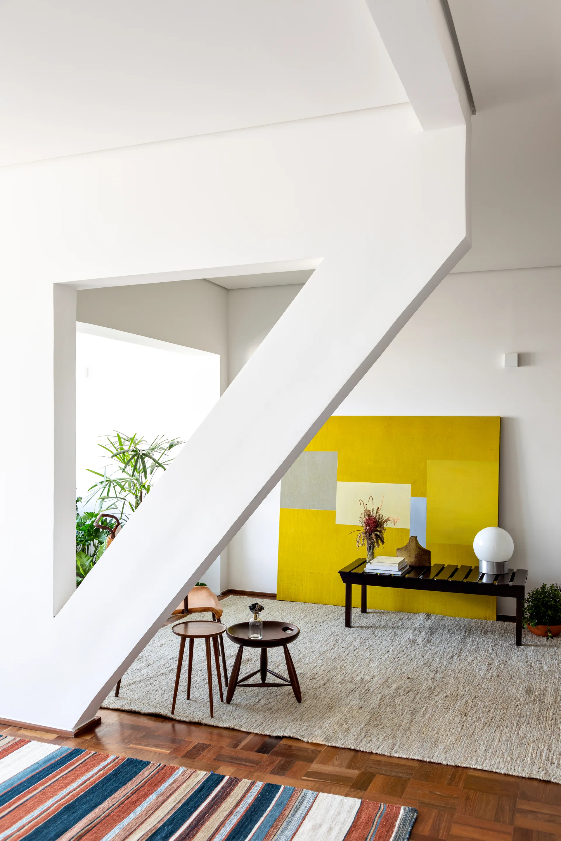 A bright modern living room featuring a large yellow abstract painting behind a black bench and an angular white staircase.