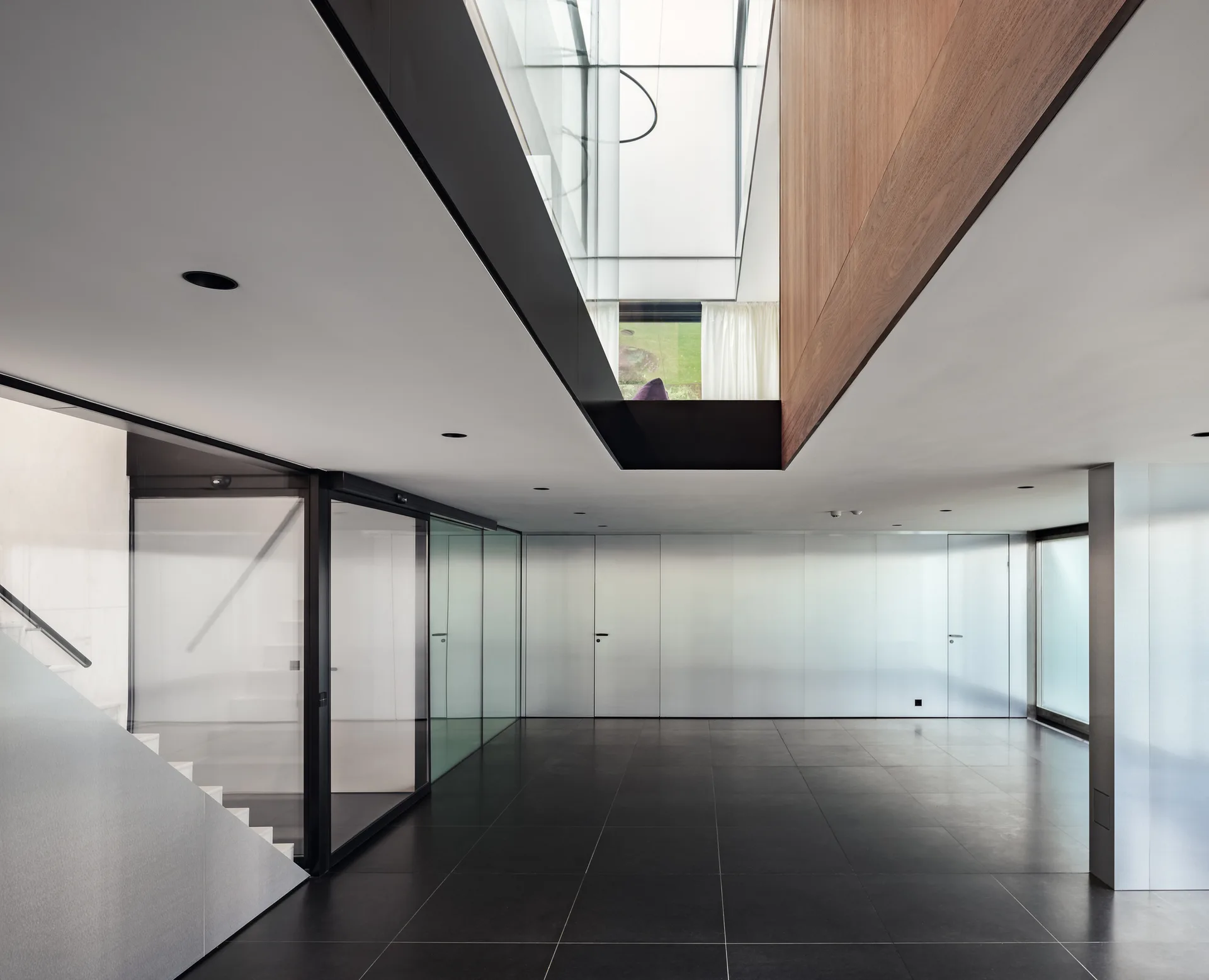 A minimalist open hallway features large dark tile flooring, glass partition walls, and a central double-height ceiling cutout with a glass skylight.