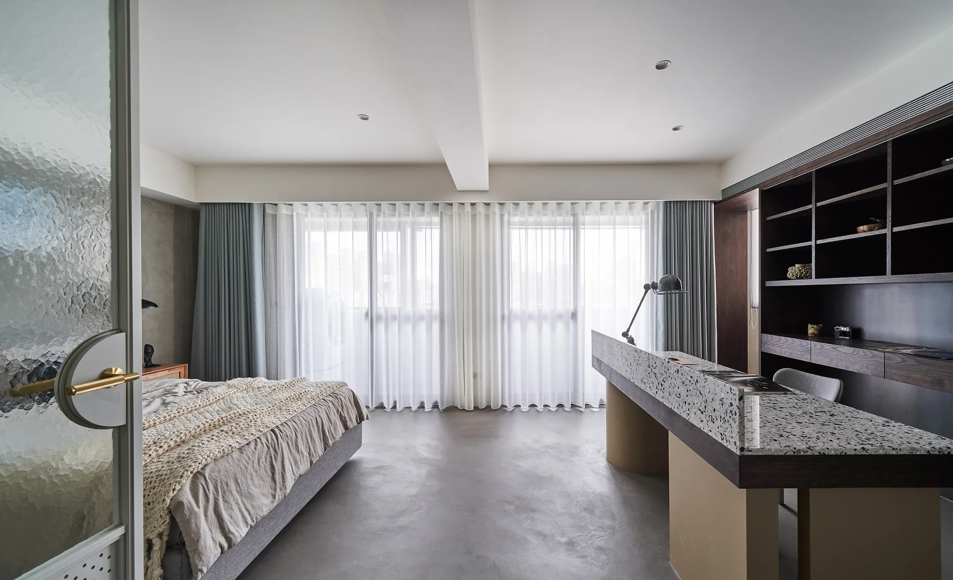A modern bedroom featuring an upholstered bed, a gray concrete floor, and a terrazzo-topped desk in front of dark wood shelving.