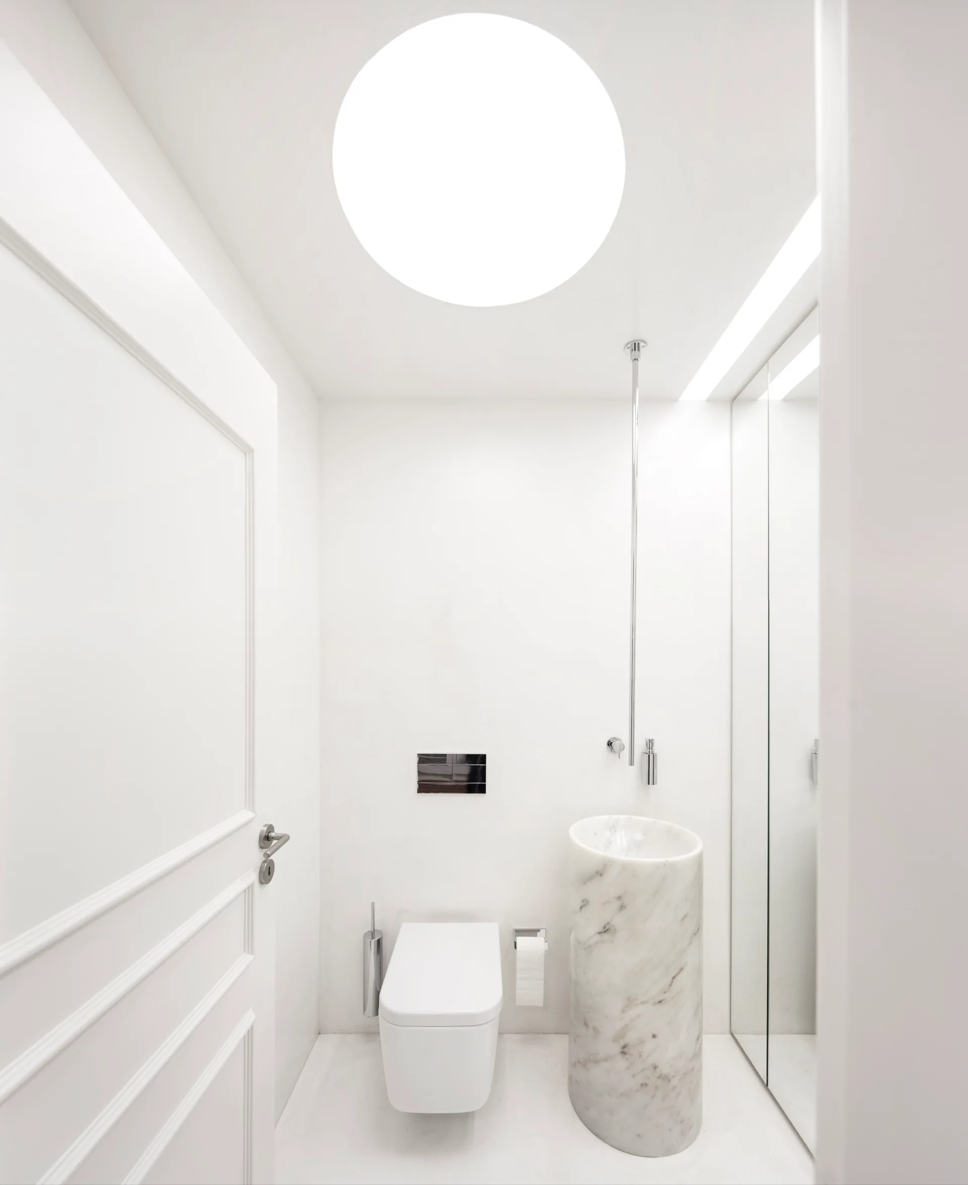 A minimalist white powder room featuring a cylindrical marble pedestal sink, a wall-mounted toilet, and a large circular ceiling light.