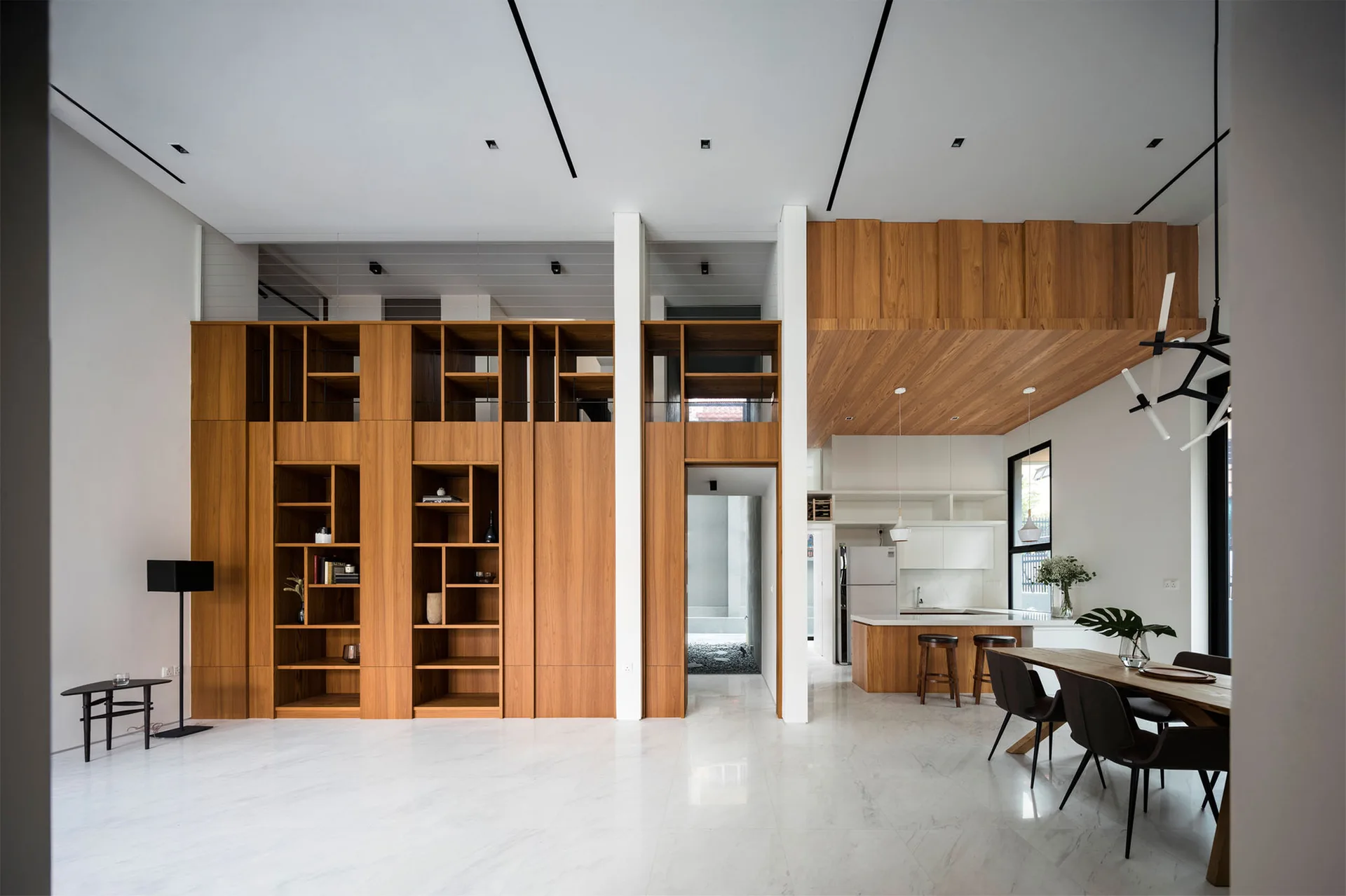 A spacious minimalist living area featuring a tall wood cabinet wall, polished white marble floors, a kitchen island, and a dining table set.
