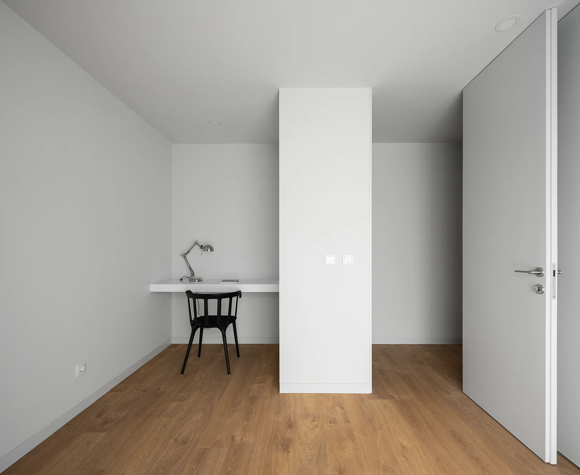 A minimalist white room featuring a floating desk and black chair set into a niche next to a central partition column on wood floors.