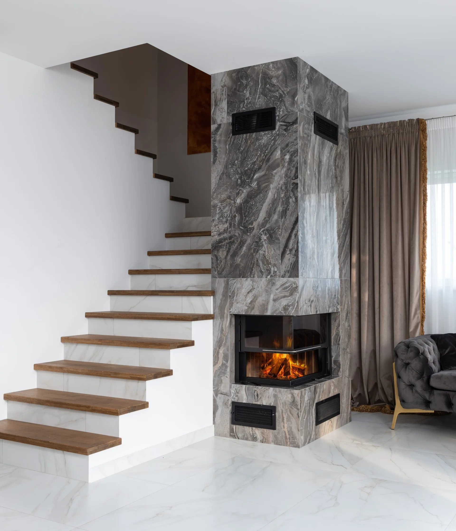 A tall grey marble corner fireplace stands next to a white staircase with wooden treads on a white tiled floor.