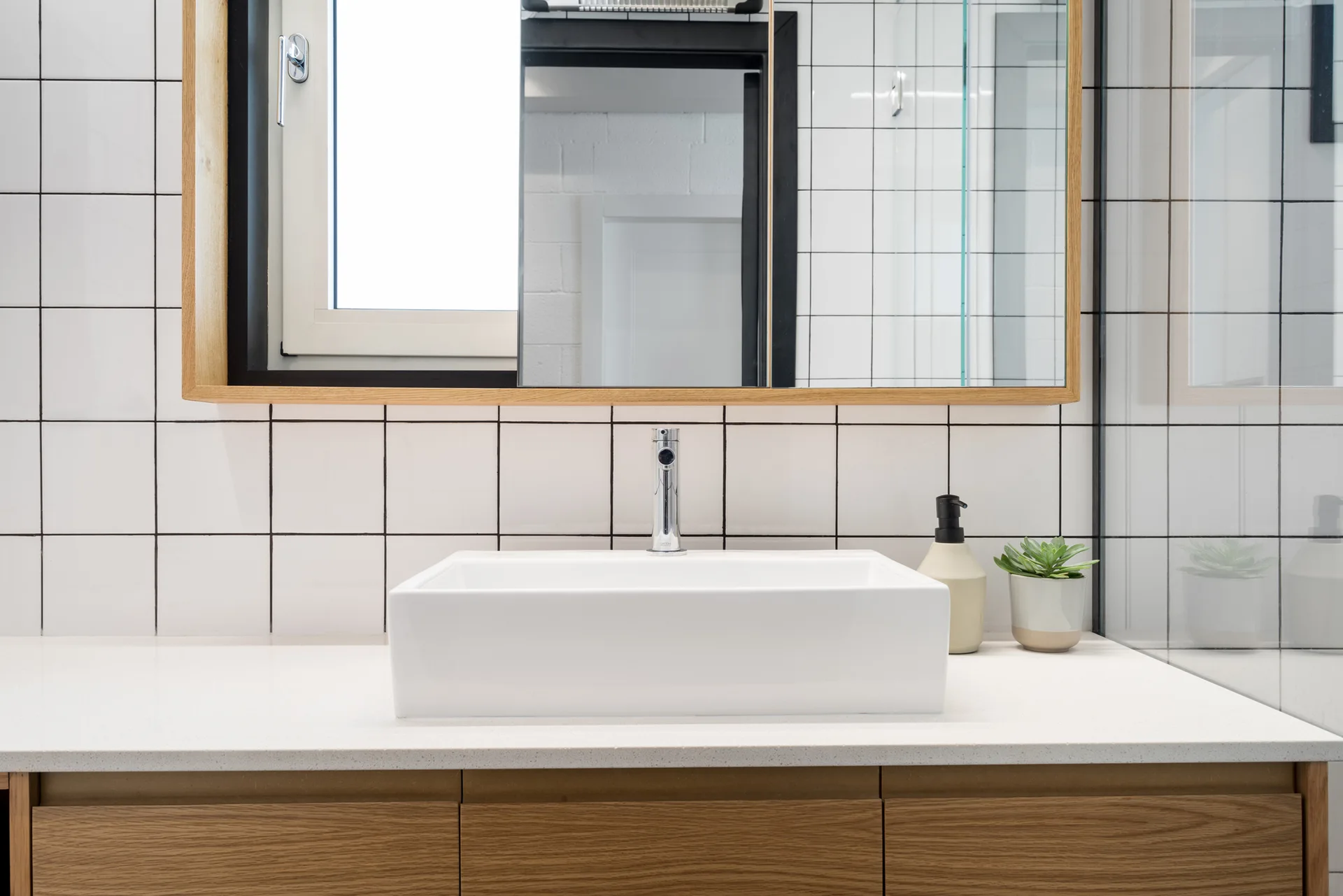 A modern bathroom vanity features a rectangular white vessel sink on a light wood cabinet against a white square-tiled wall with dark grout.