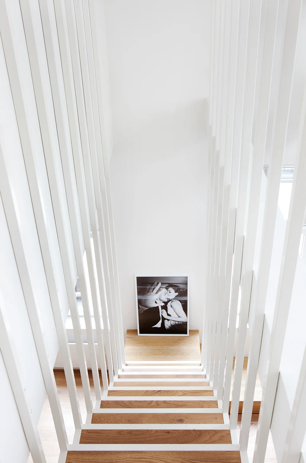 Looking down a modern staircase with light wood treads, vertical white slatted railings, and a black-and-white photograph resting on the landing.