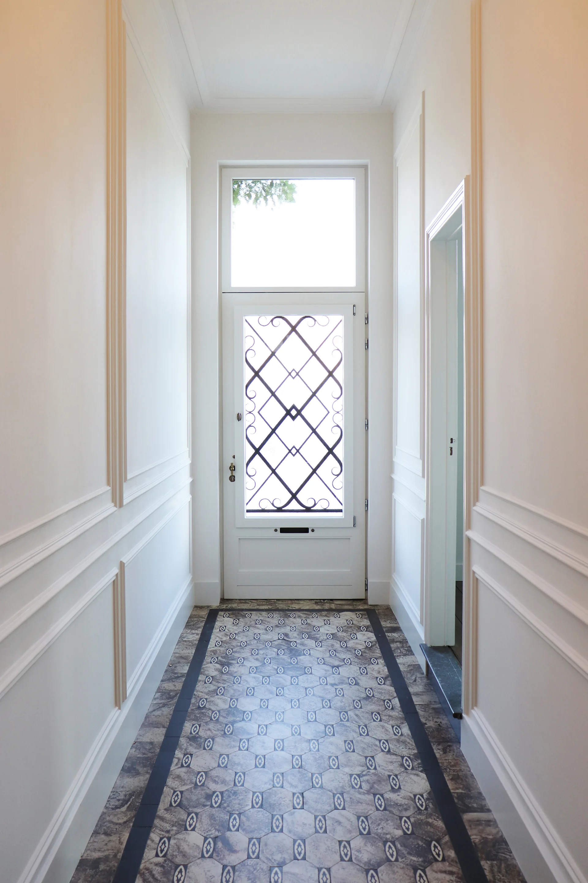 A bright white hallway with decorative wall molding features a geometric patterned tile floor leading to a door with a black decorative iron grille.