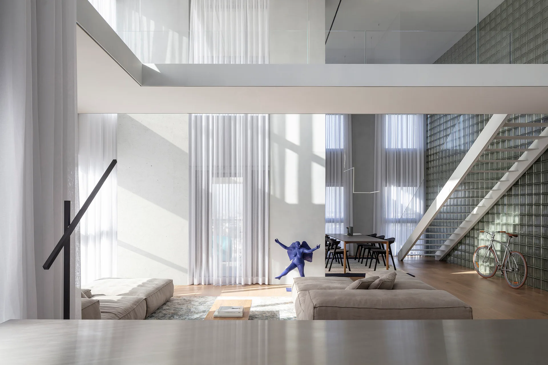 A double-height modern living room features a tall glass block wall, low-profile beige sectional sofas, white sheer curtains, and a mezzanine balcony.