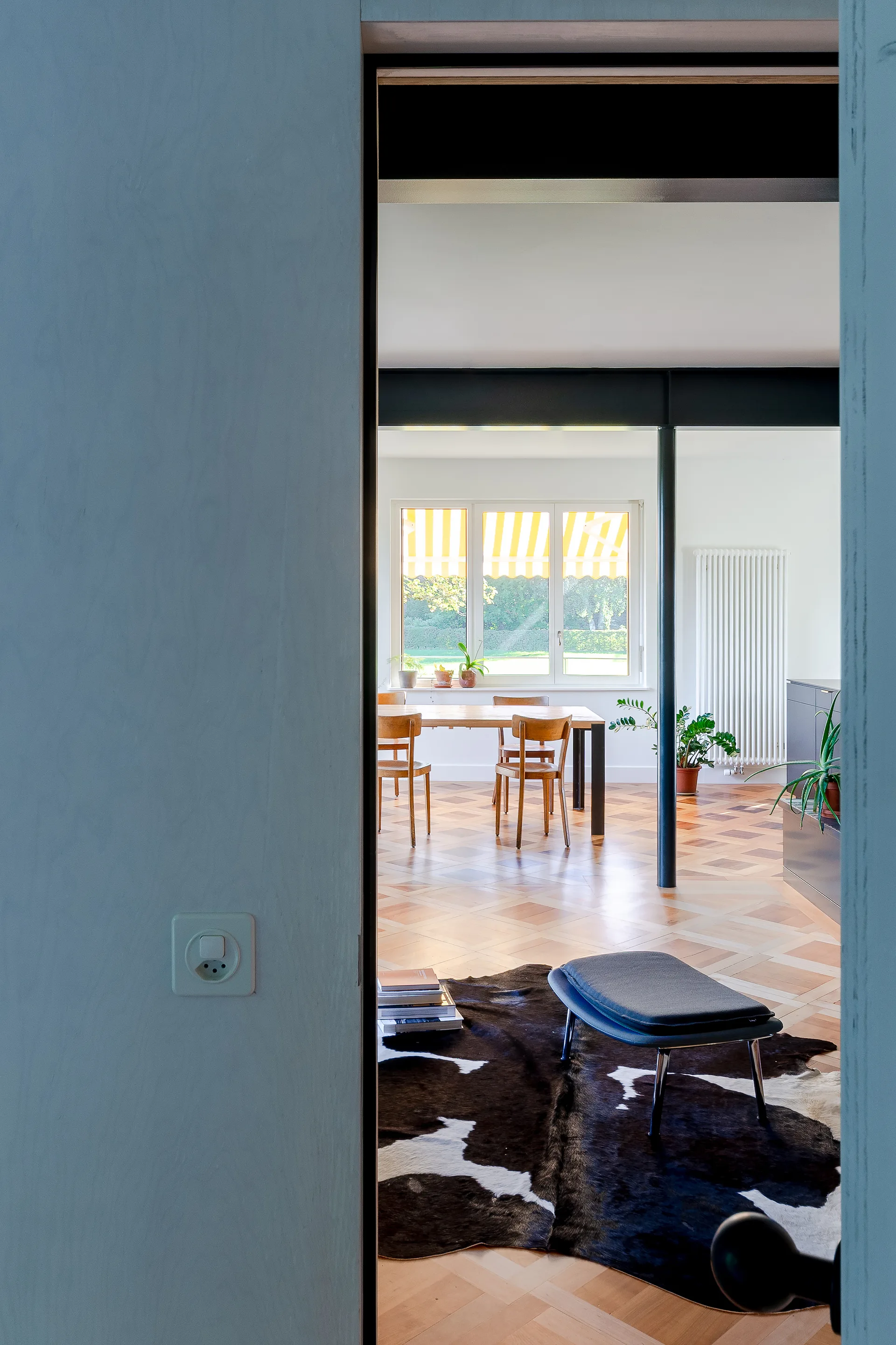 A view through a white-walled doorway reveals a bright dining room with geometric parquet flooring, a wooden table, and a dark structural column.