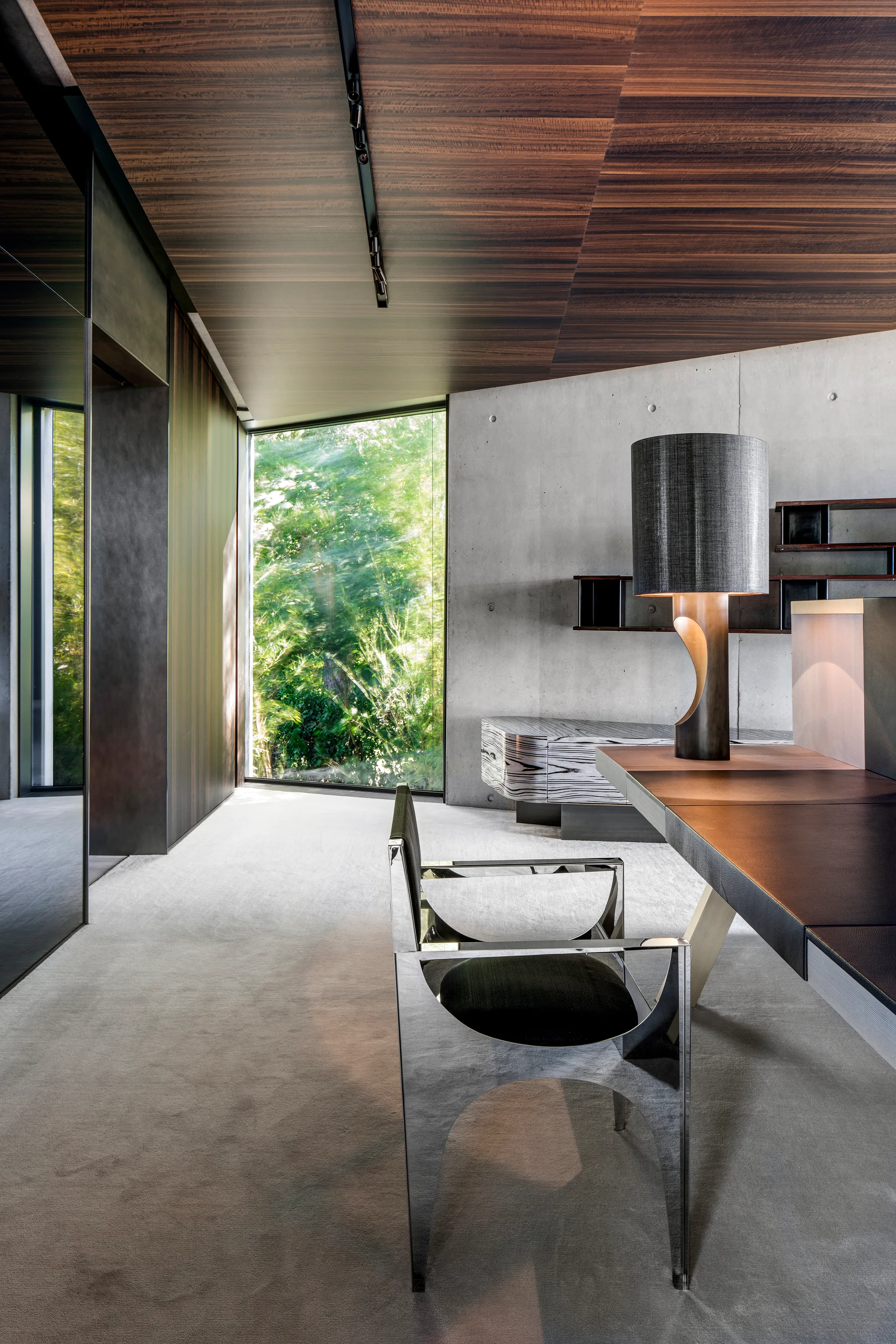 A modern home office features a polished metal chair, concrete walls, a dark desk, and a warm wood-paneled ceiling.
