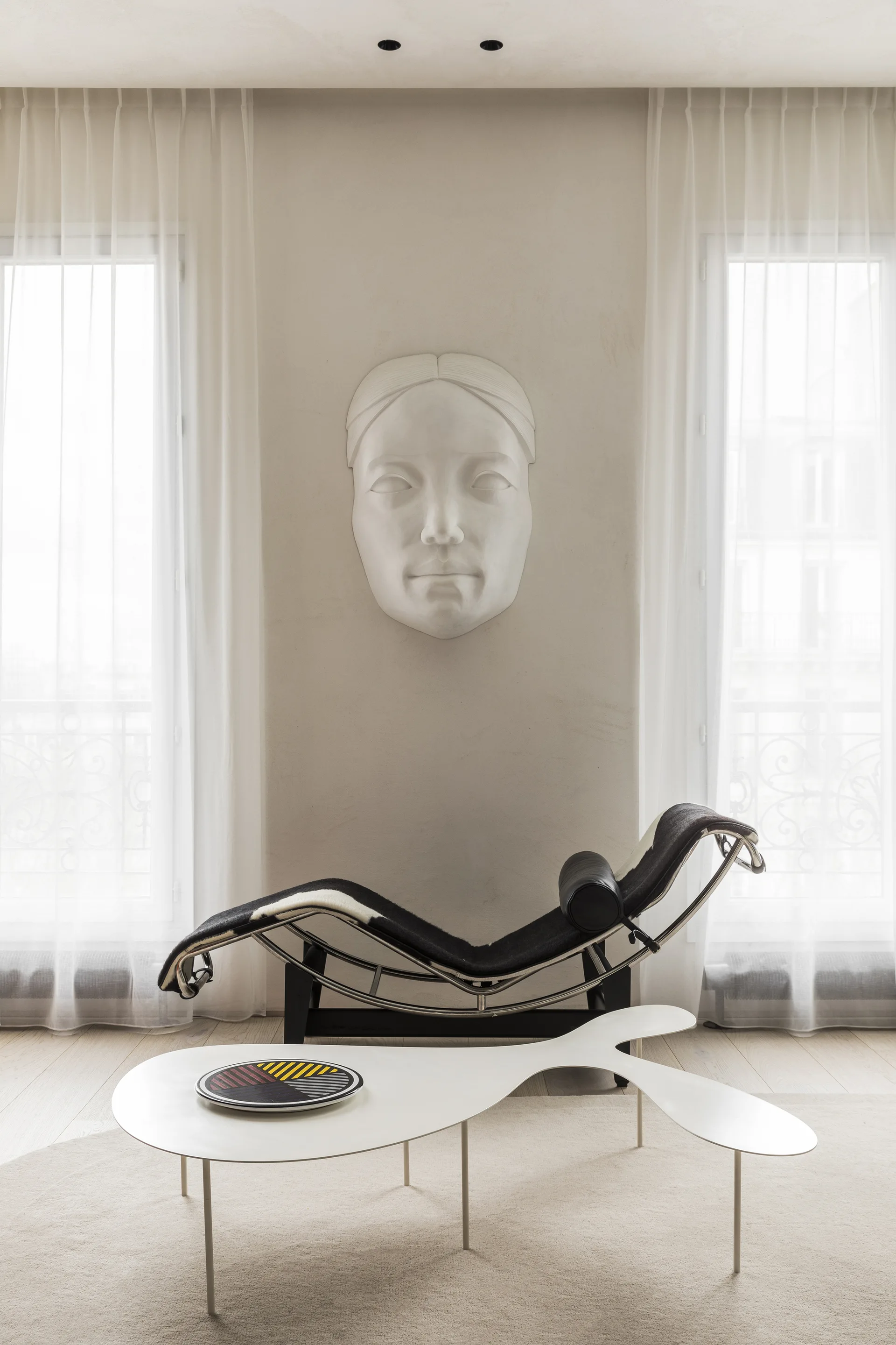 A modernist black and white chaise lounge placed between two draped windows, with a large white plaster face sculpture mounted on the wall above.