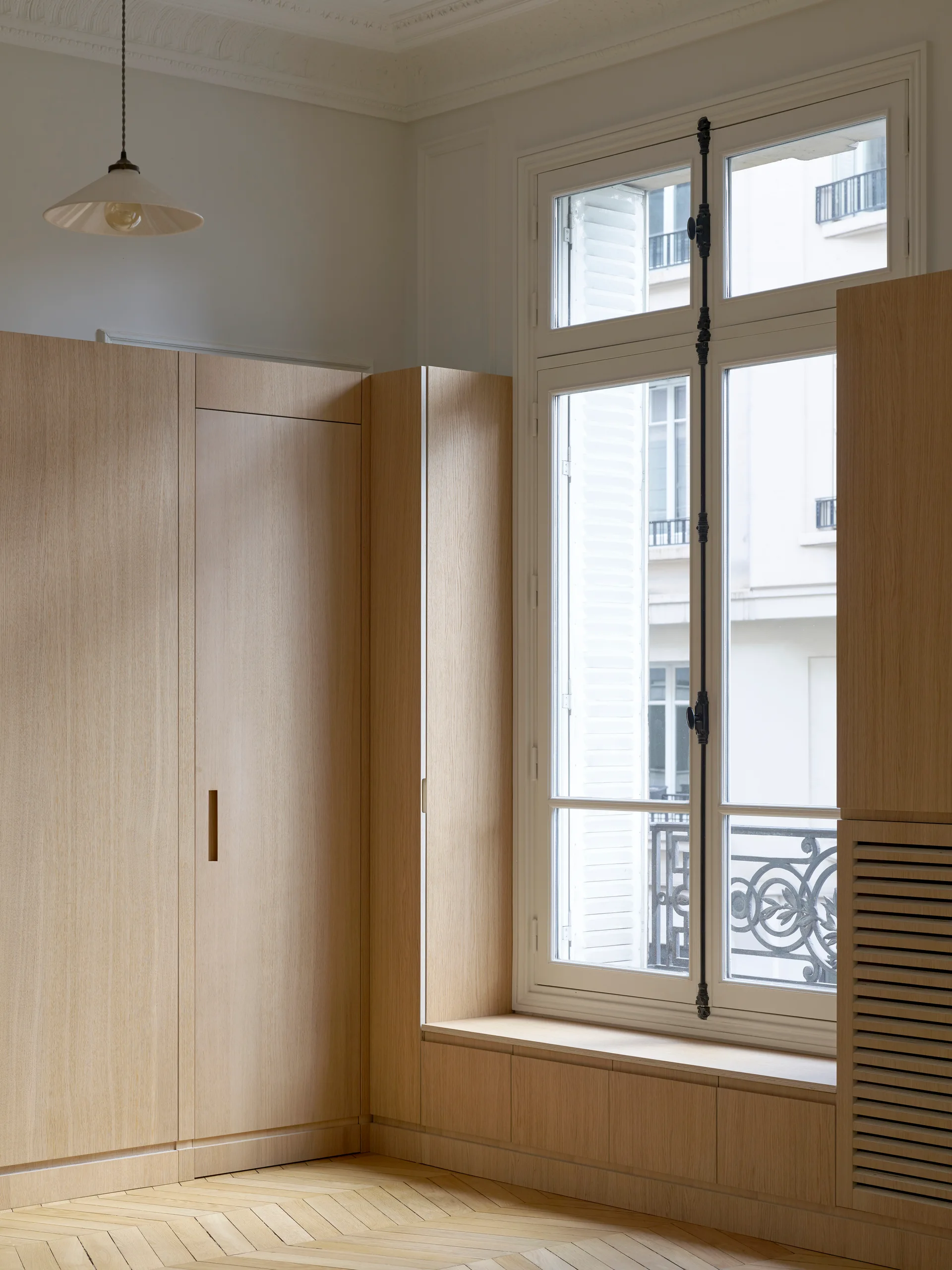 Light oak built-in cabinetry frames a tall white window above herringbone pattern parquet floors.