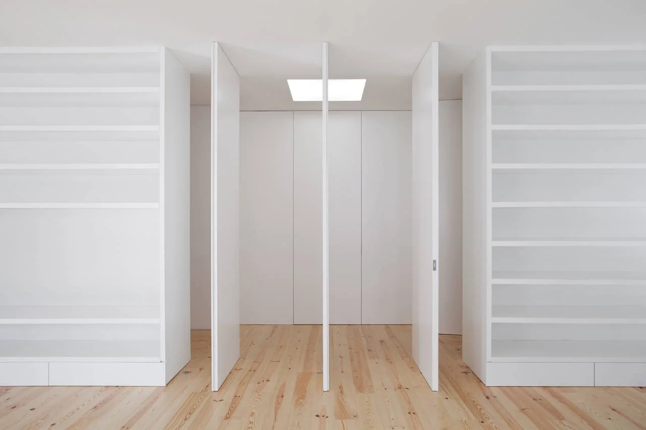 Symmetrical white minimalist closet featuring built-in shelving, pivoting center doors, light wood flooring, and a square ceiling skylight.
