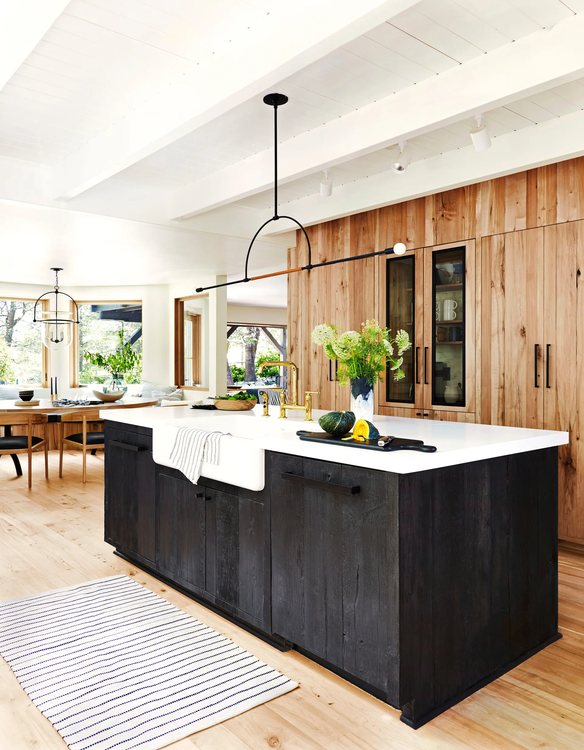 A bright kitchen features a black island with a white farmhouse sink, gold faucet, and a wall of natural wood cabinetry.