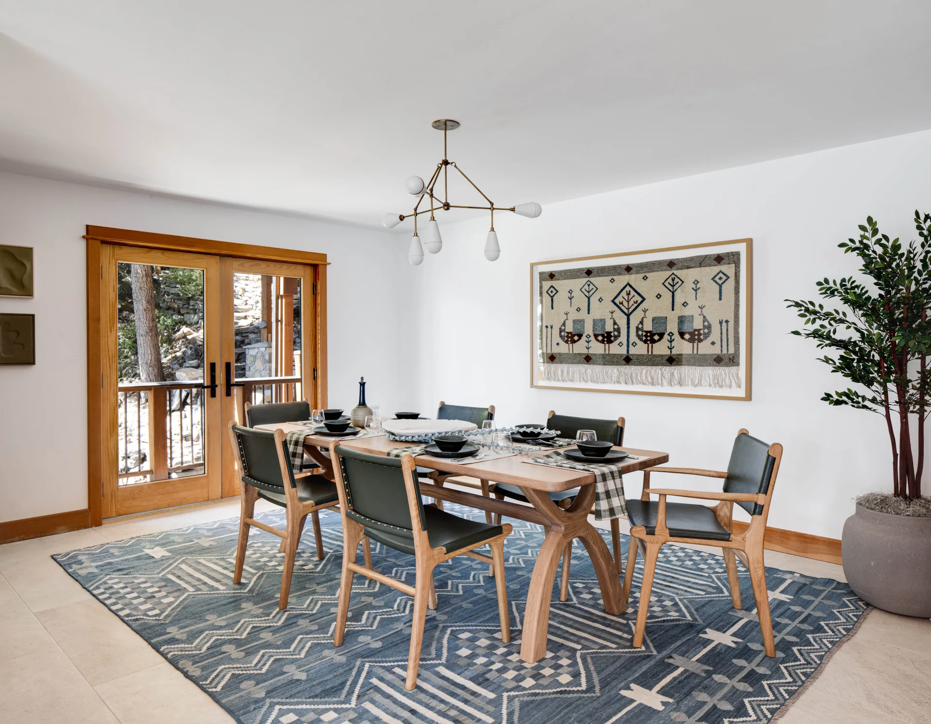 A wooden dining table and green-seated chairs sit on a blue patterned rug in a bright dining room with wood-trimmed glass doors.