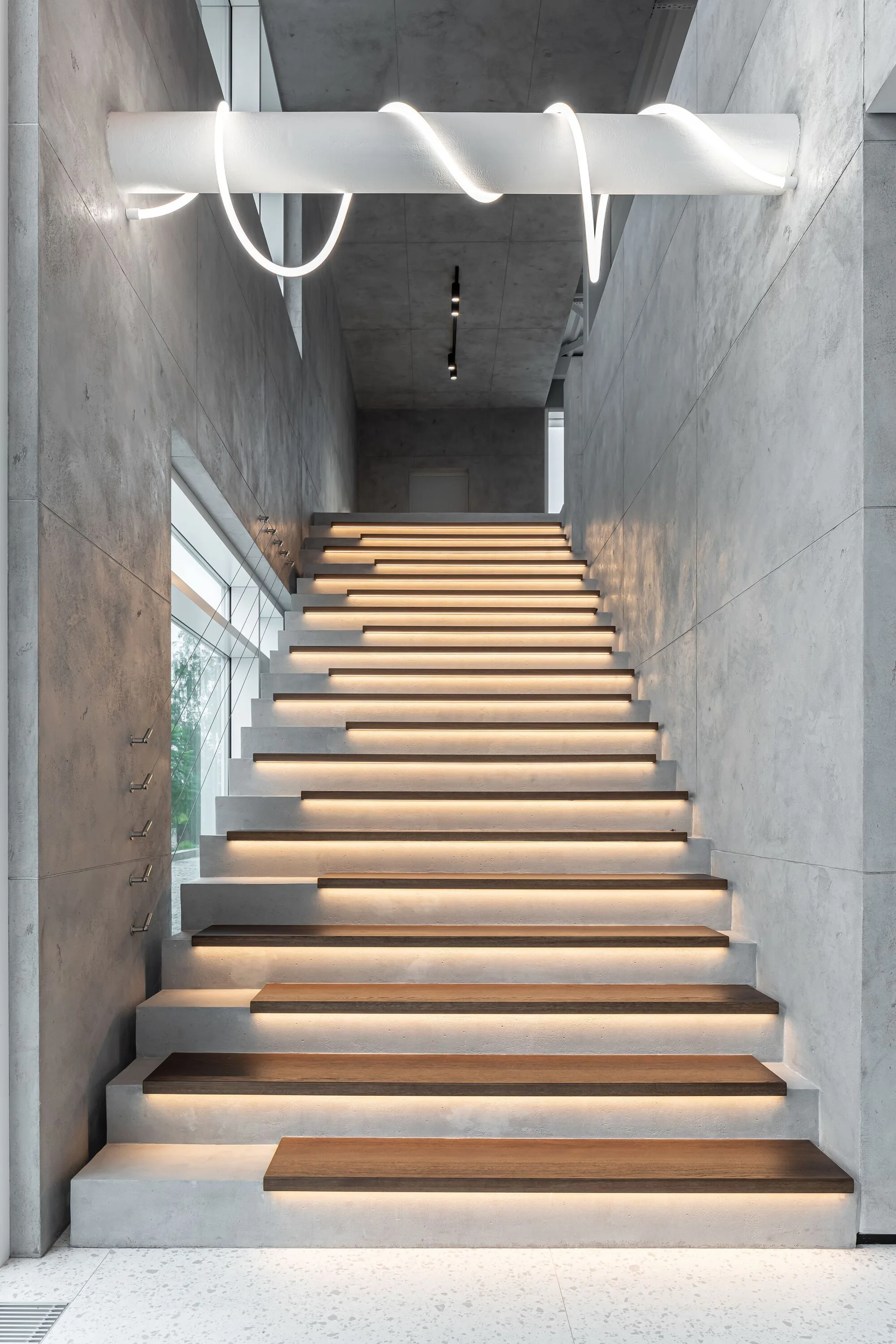 A modern staircase with concrete walls features staggered wooden treads illuminated by integrated LED strip lighting under a sculptural white overhead light.