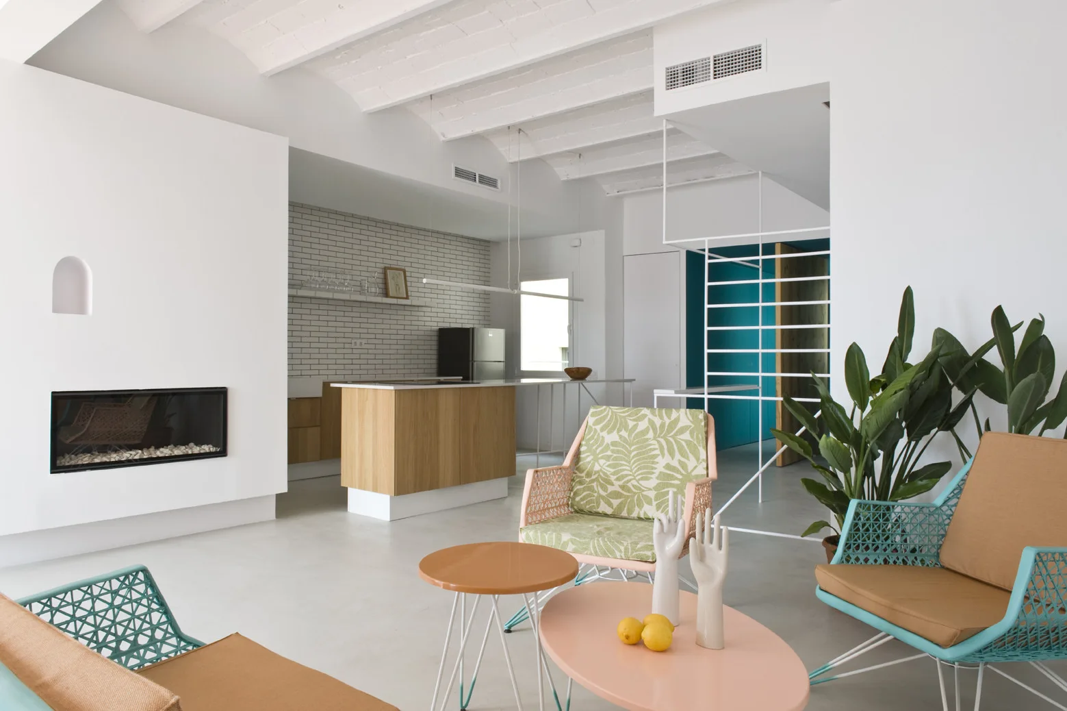 Pastel geometric armchairs sit in a white living room with a fireplace, open kitchen, and exposed ceiling beams.