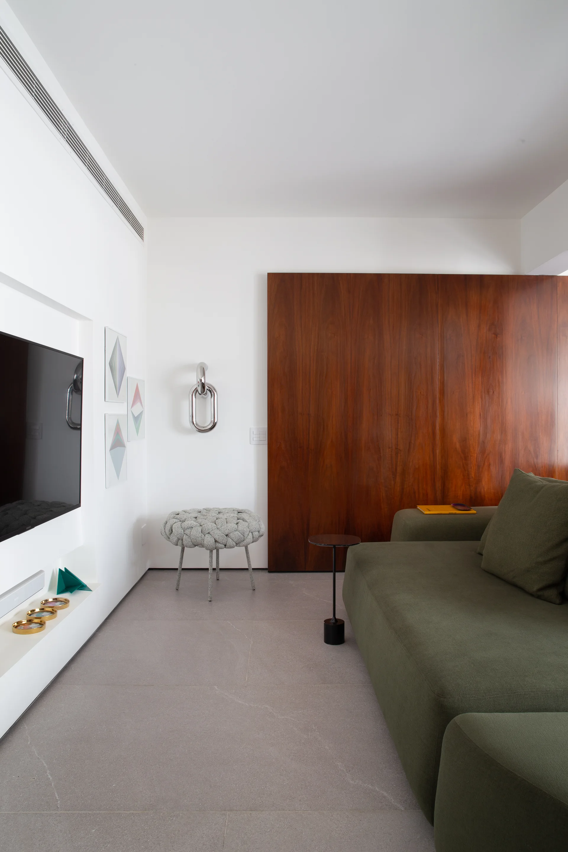 An olive green sofa and a textured grey knit ottoman sit on stone tile flooring opposite a wall-mounted TV and a dark wood accent panel.
