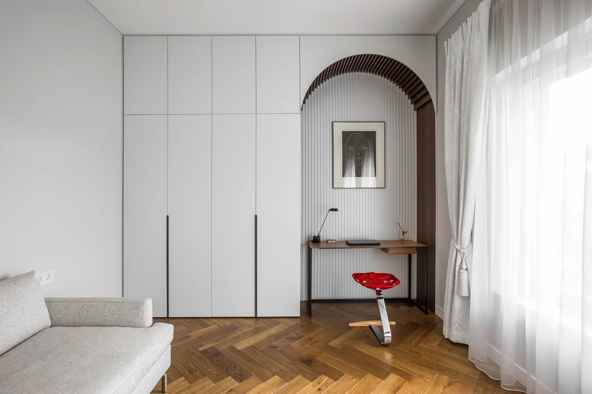 A minimalist bedroom featuring white built-in wardrobes, an arched desk alcove with a red stool, and herringbone wood floors.