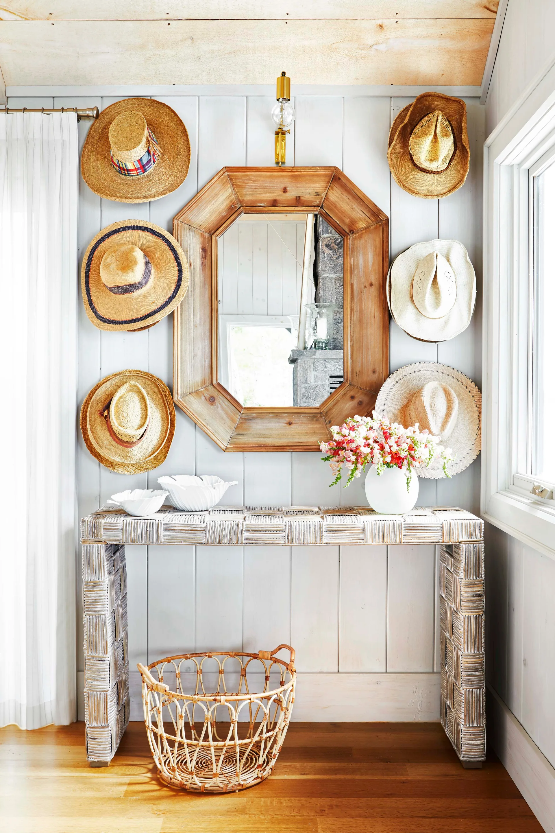 An octagonal wood-framed mirror is flanked by six straw hats above a woven console table and a wicker basket on a hardwood floor.