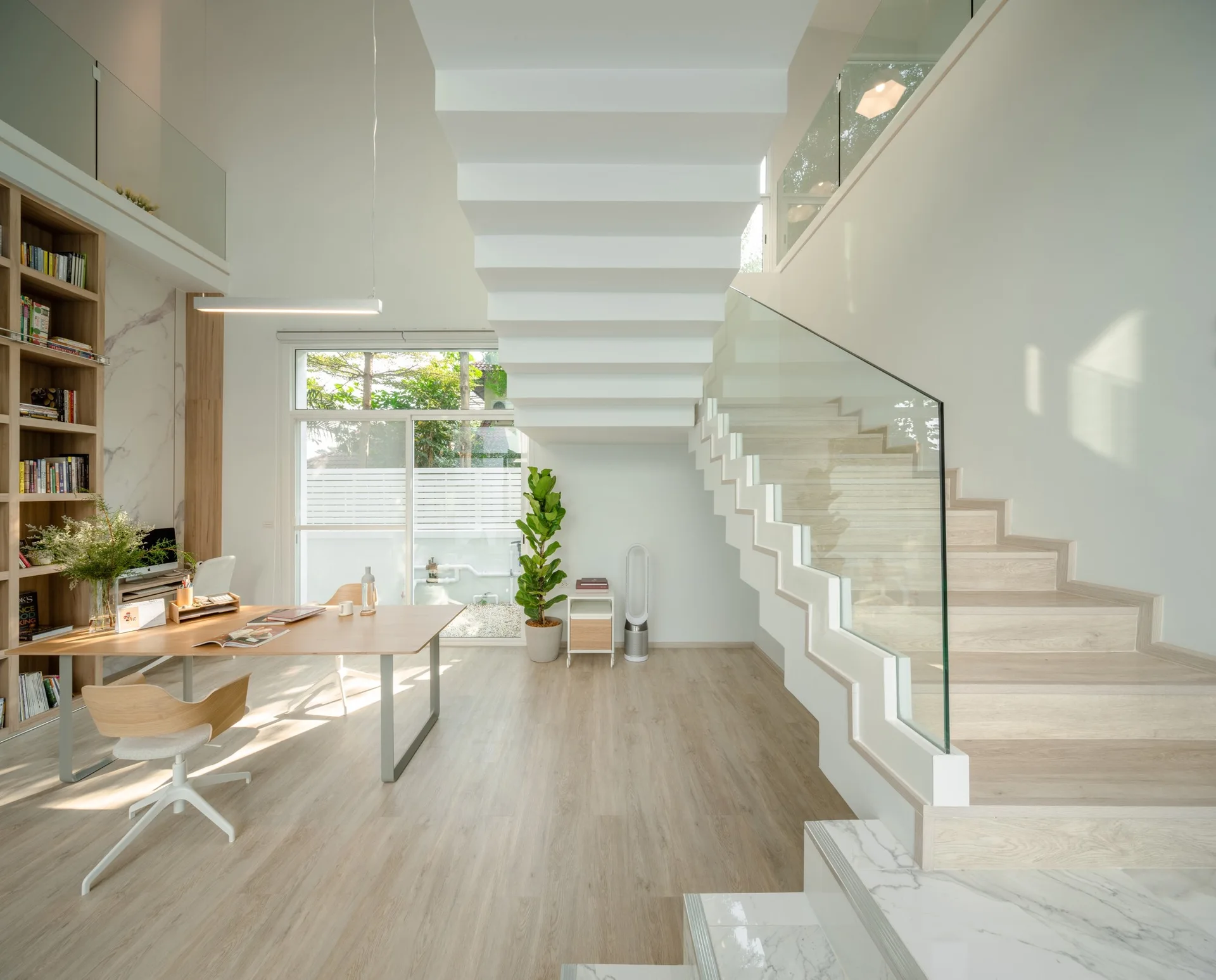 A bright home office features a wooden desk, a floor-to-ceiling built-in bookshelf, and a white modern staircase with glass railings.