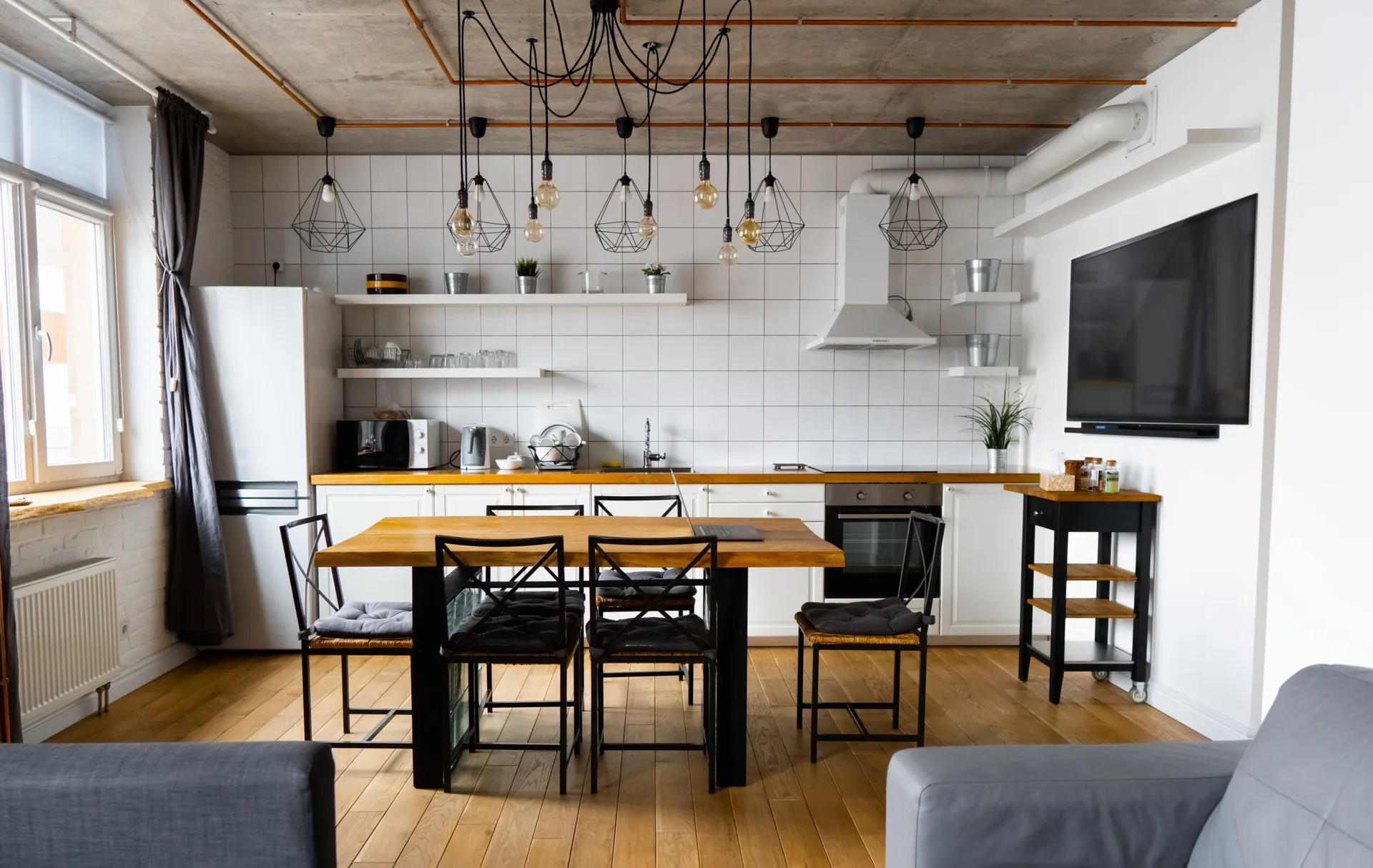 An industrial kitchen features white square tile walls, a concrete ceiling with exposed pipes, and a wooden dining table with black chairs.