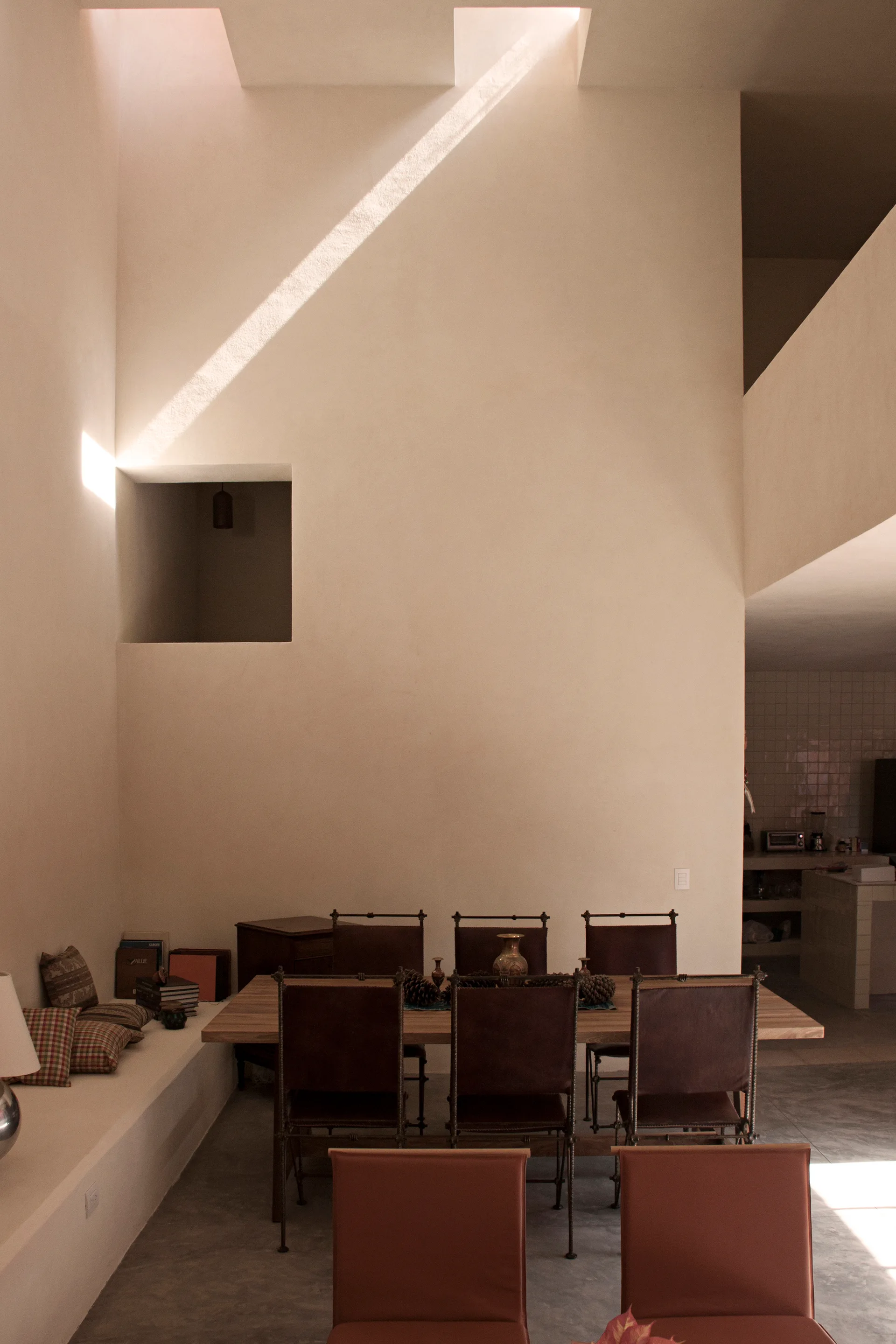 A minimalist dining room features a wooden table with leather chairs set against a tall cream plaster wall illuminated by a beam of light from a skylight.