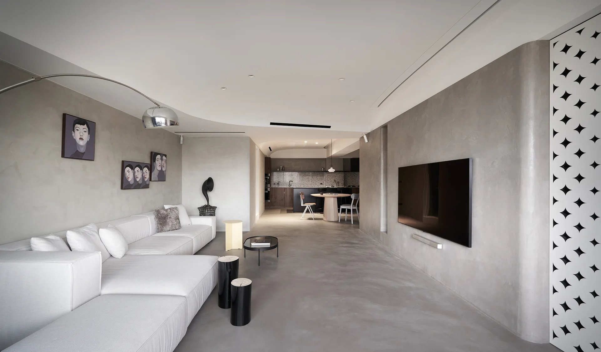 A minimalist living room features grey plaster walls and floors, a large white sectional sofa, a wall-mounted television, and a curved ceiling.