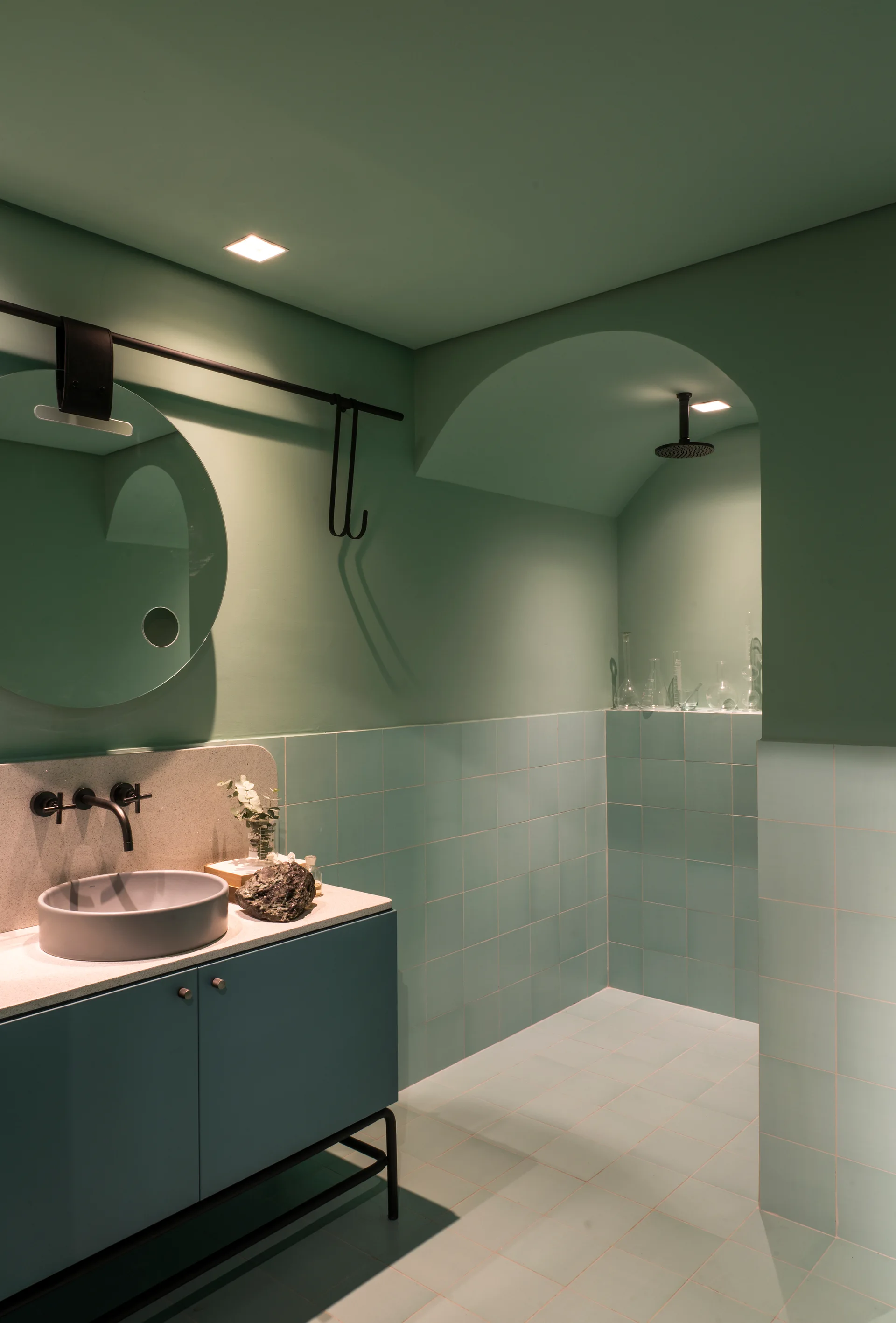 A modern mint green bathroom featuring a teal tiled lower wall, an arched shower entrance, and a teal vanity with a round mirror.