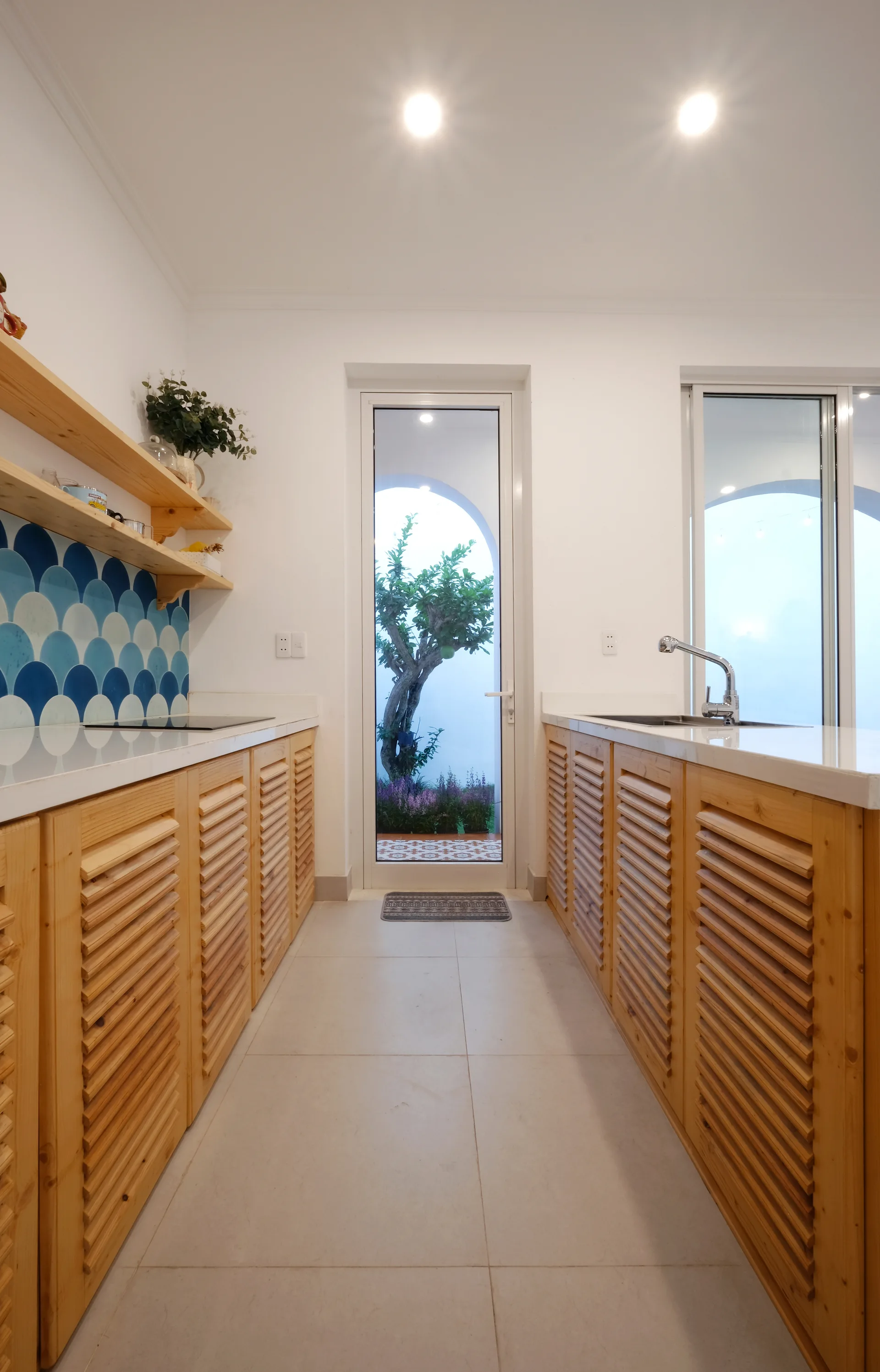 A galley kitchen features light wood louvered cabinets, white countertops, a blue scalloped tile backsplash, and a glass door leading to a garden.