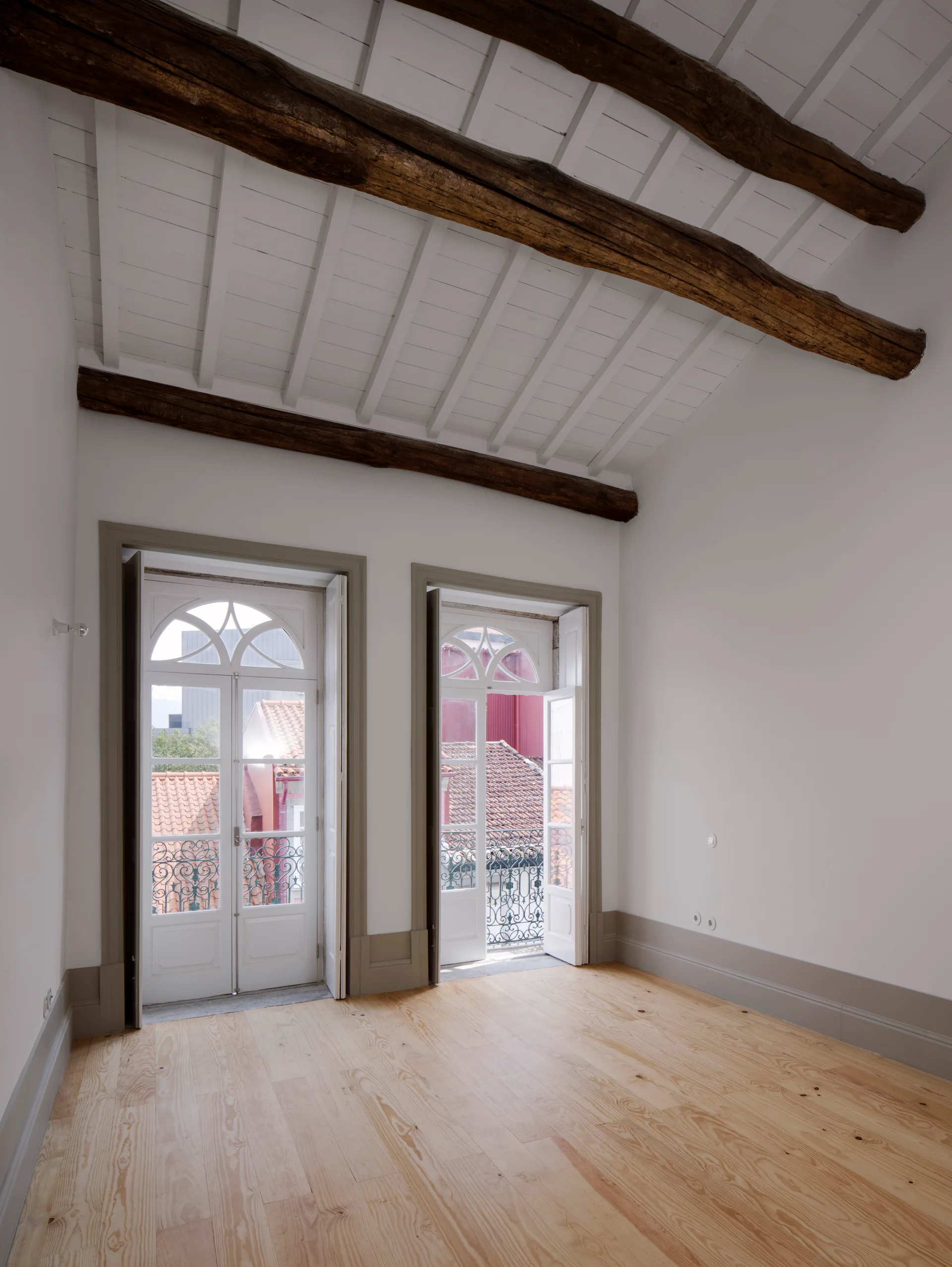 An empty room with exposed dark wood ceiling beams, light wood floors, and two arched French doors framed in taupe-painted trim.