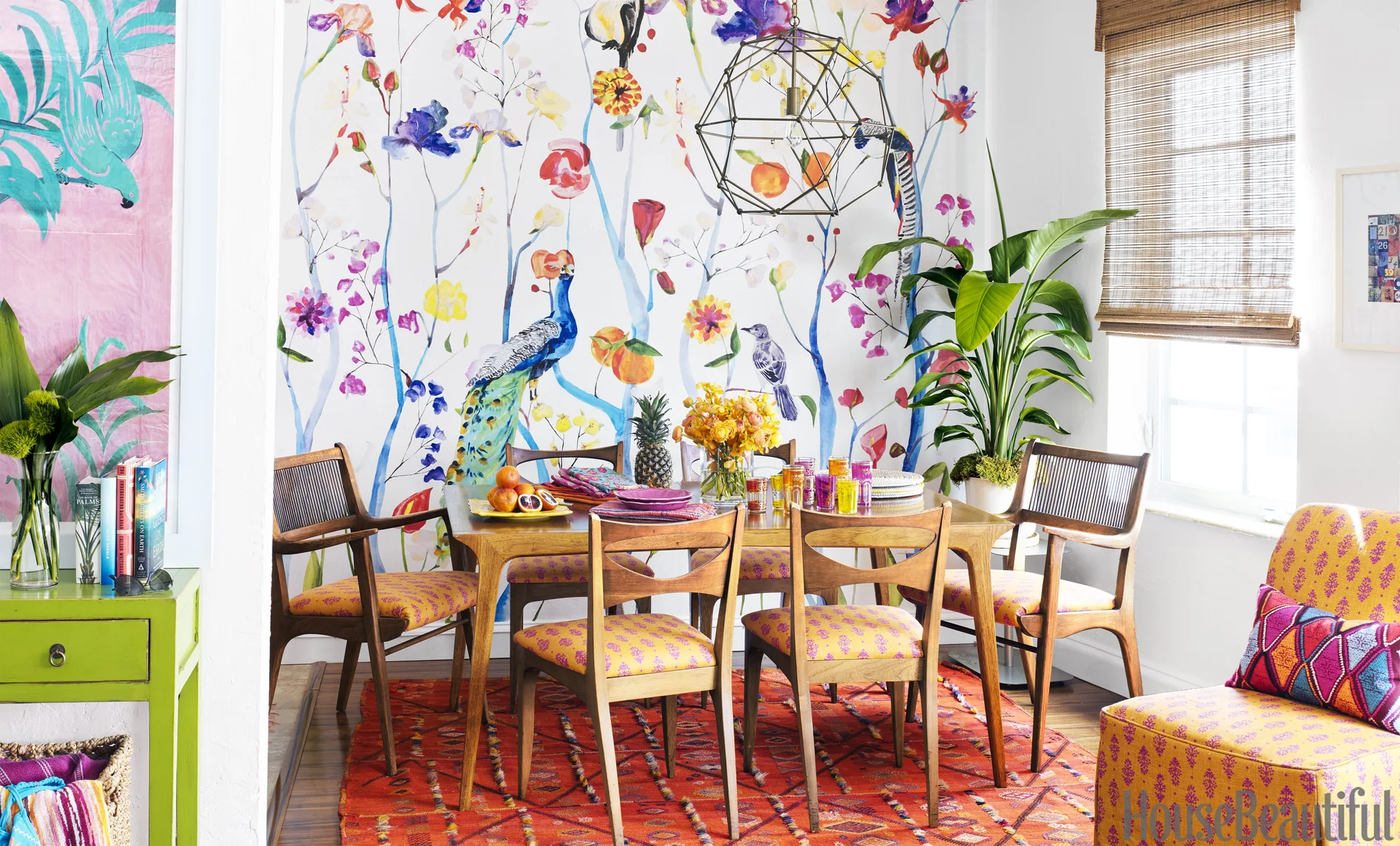 A mid-century modern wooden dining set sits on an orange patterned rug in front of a colorful floral wallpaper mural with birds.