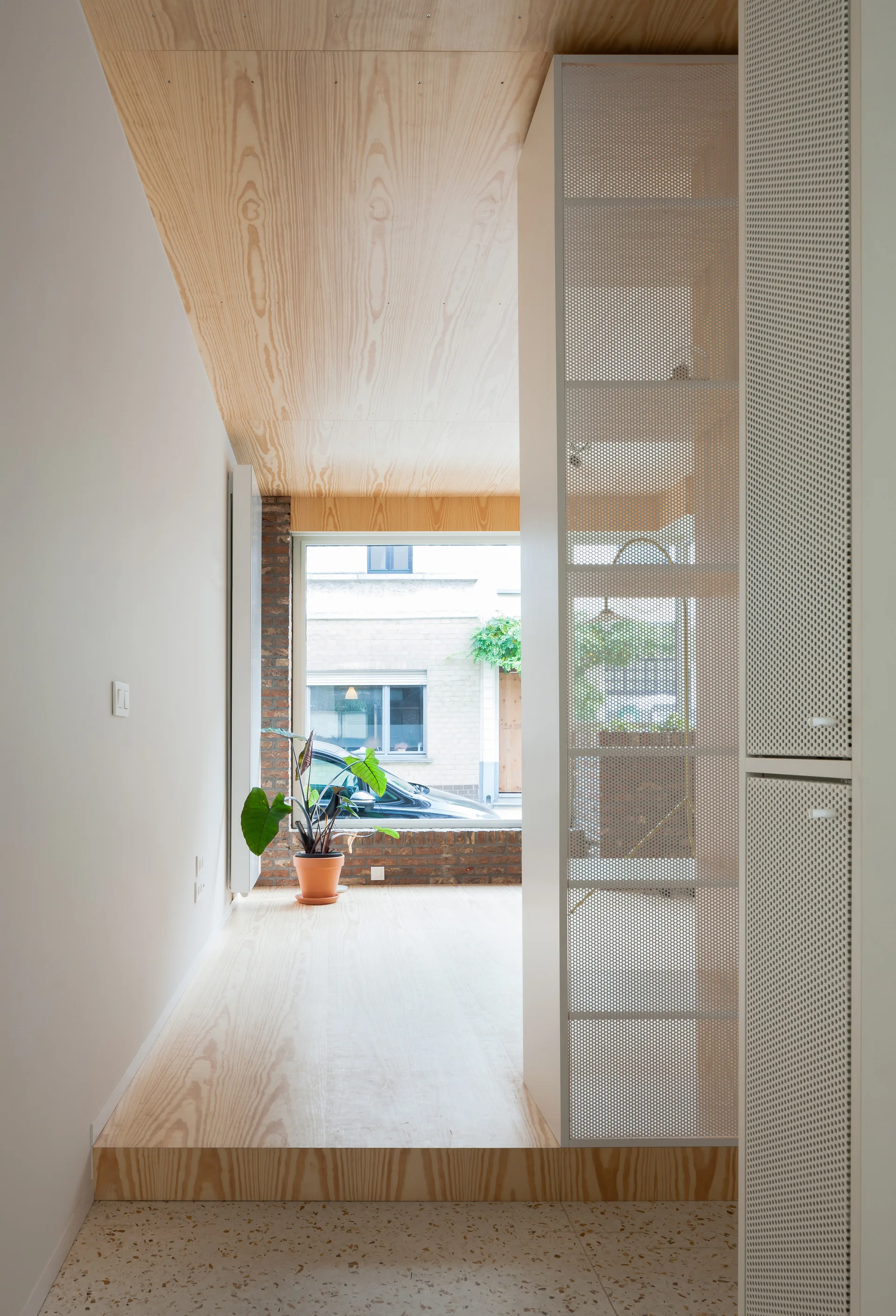 A modern entryway features a speckled terrazzo floor stepping up to a raised plywood platform next to a white perforated metal room divider.
