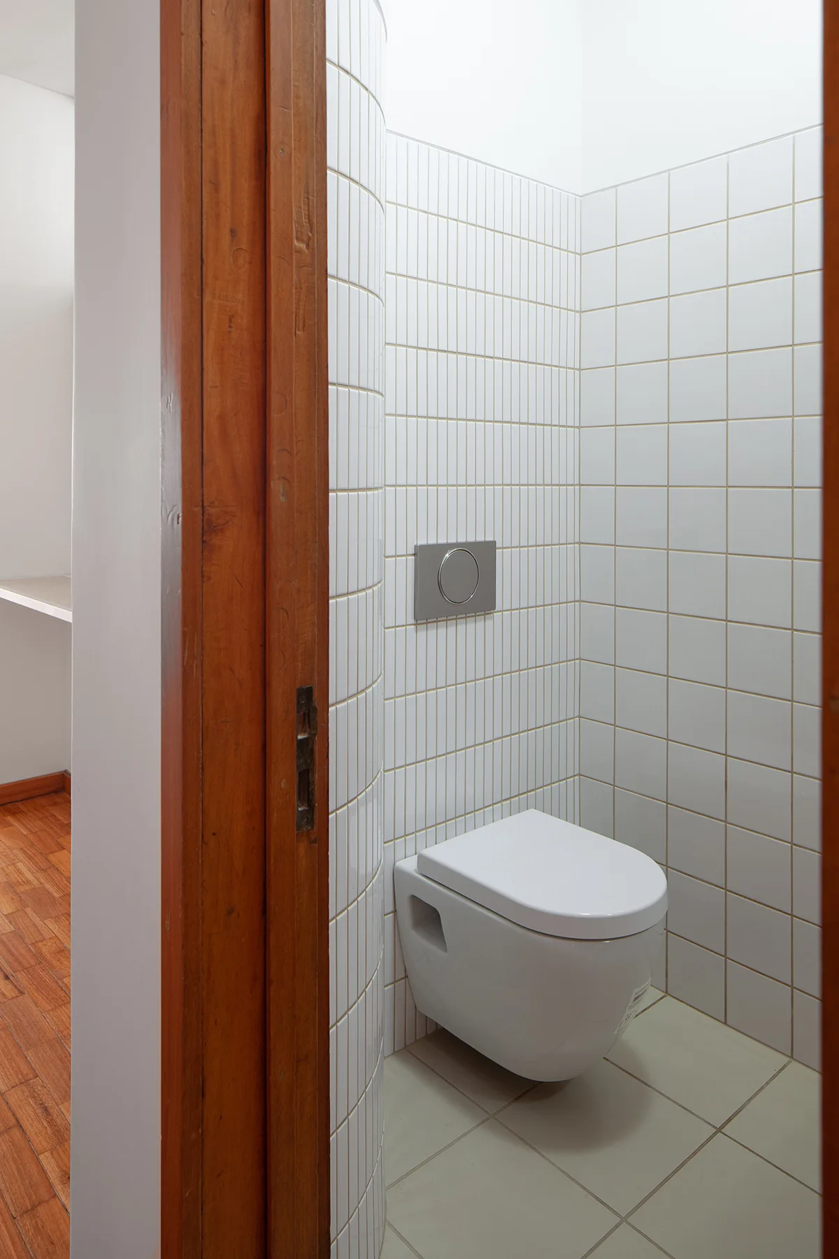 A minimalist powder room features a white wall-hung toilet, square white tiled walls, a curved tiled partition, and a warm wood door frame.